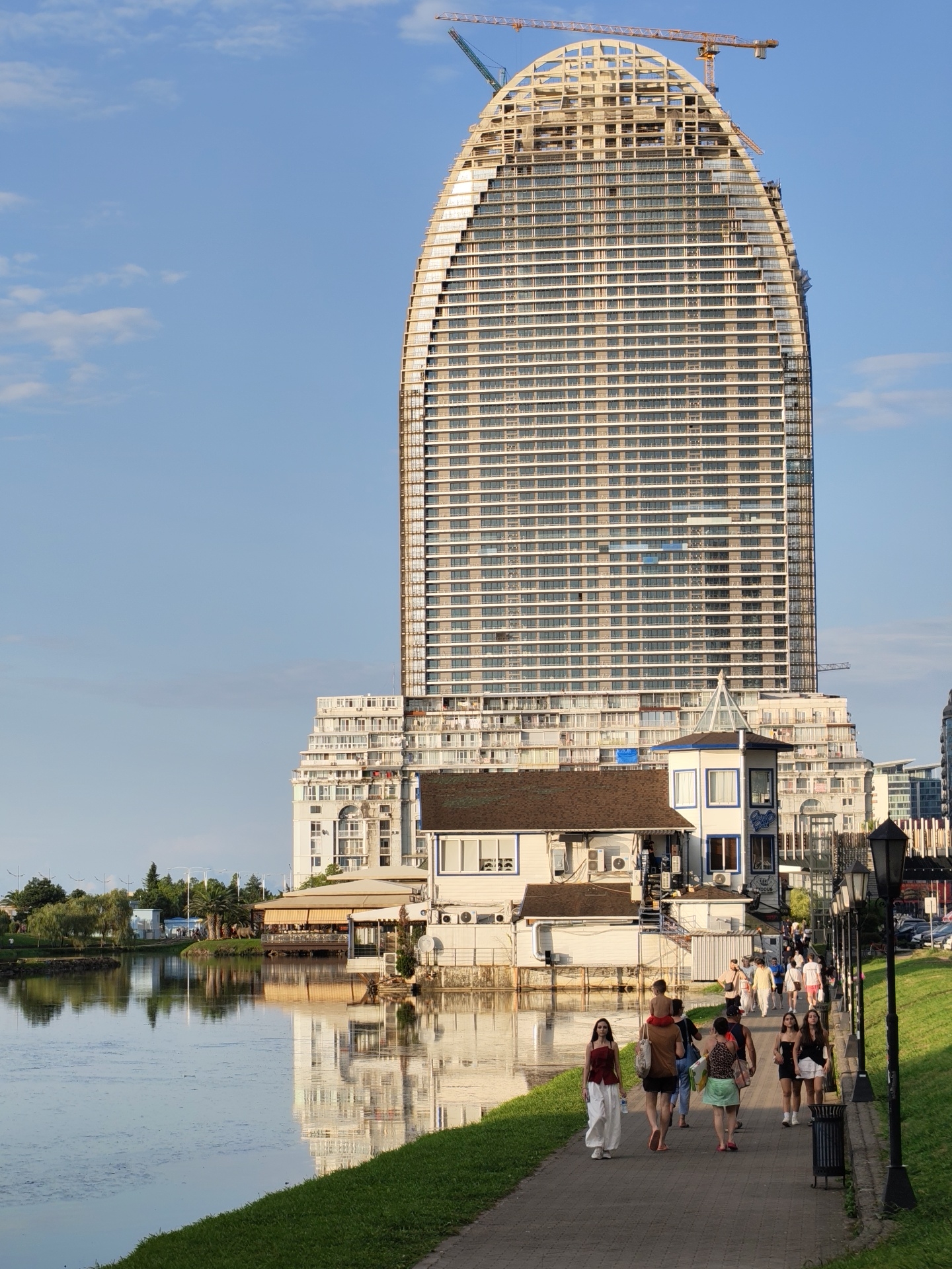 The image depicts a sunny afternoon in Batumi, Georgia, likely along the Batumi Boulevard. In the foreground, a paved walkway runs alongside a calm body of water, which reflects the surrounding buildings and the sky. A strip of green grass borders the walkway on one side, and on the other side, adjacent to the water, are several low-rise buildings, including a restaurant with a yellow awning. Further back, dominating the scene, is a very tall, partially constructed building with a distinctive curved, egg-like shape. This skyscraper is covered in scaffolding and is clearly under construction, with cranes visible at the top. People are walking along the paved path, some individually, others in small groups, enjoying the promenade. The sky is a clear blue with a few wispy clouds, and the overall atmosphere suggests a pleasant day in a coastal city.