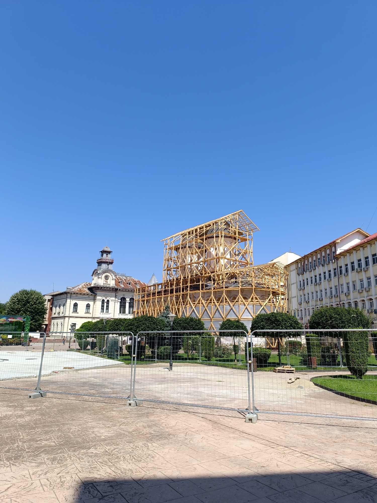 In Târgu Jiu, Romania, a construction scene unfolds under a clear blue sky. A large building, possibly a historical landmark, is covered in a temporary wooden scaffolding, suggesting ongoing restoration or renovation work. The scaffolding forms a geometric pattern, enveloping the upper portion of the structure. To the left of the scaffolding, a distinct building with a clock tower stands out, featuring a rusted roof and ornate architectural details. Further to the right, a modern, multi-story building with numerous windows and air conditioning units is visible.
The foreground is dominated by a paved area with a textured pattern, likely a public square or walkway. Several metal barriers, commonly used for crowd control or to demarcate construction zones, are erected across the scene. Behind the barriers, a manicured green space with well-trimmed bushes and trees provides a contrast to the construction. The presence of the scaffolding and barriers clearly indicates an active construction or repair project underway in this urban environment. No people are readily visible in the immediate vicinity of the construction.