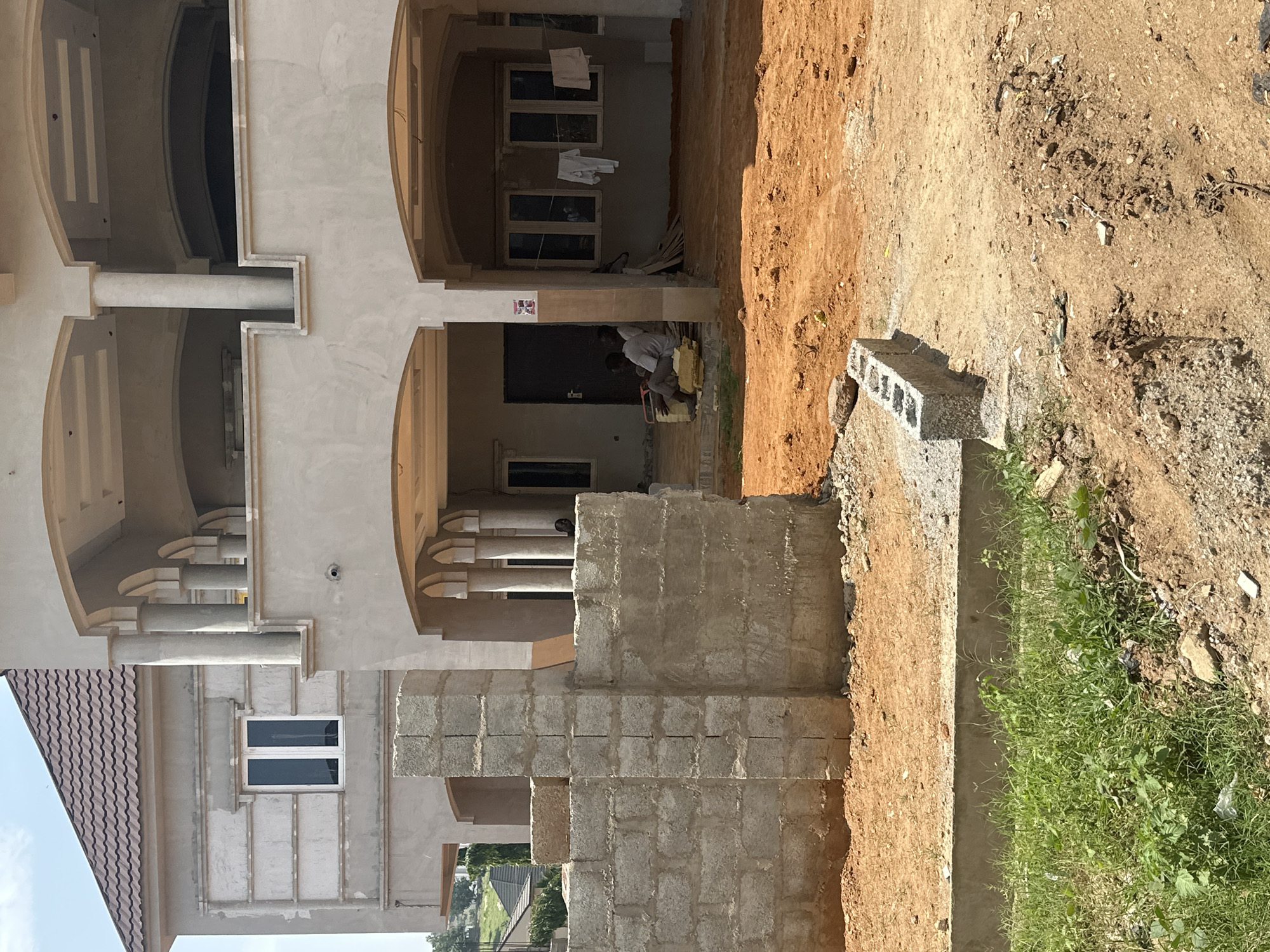 In Gwarinpa, Nigeria, a construction site is captured under a bright sky. The foreground features an area of dirt with some overgrown grass and scattered debris, including a small section of a concrete border and a few loose concrete blocks. To the left, a partially constructed wall made of grey cinder blocks stands prominently. Behind this wall, a more substantial building is visible, its exterior walls are plastered and painted in a light, neutral tone. This building has a tiled roof and features several windows, including a large, double-paned window on the left side.
The central and right portion of the image shows a multi-story structure, also under construction, with a light-colored, plastered facade and arched openings on the ground floor. This building has a covered porch area supported by thick, round columns. Inside the covered area, several windows are visible, and a clothesline with white clothing hanging on it is strung across. Two men are seated on yellow blocks within this covered space. The ground in front of this structure is a reddish-orange dirt. In the background, another building with a similar tiled roof is partially visible on a slight incline.
