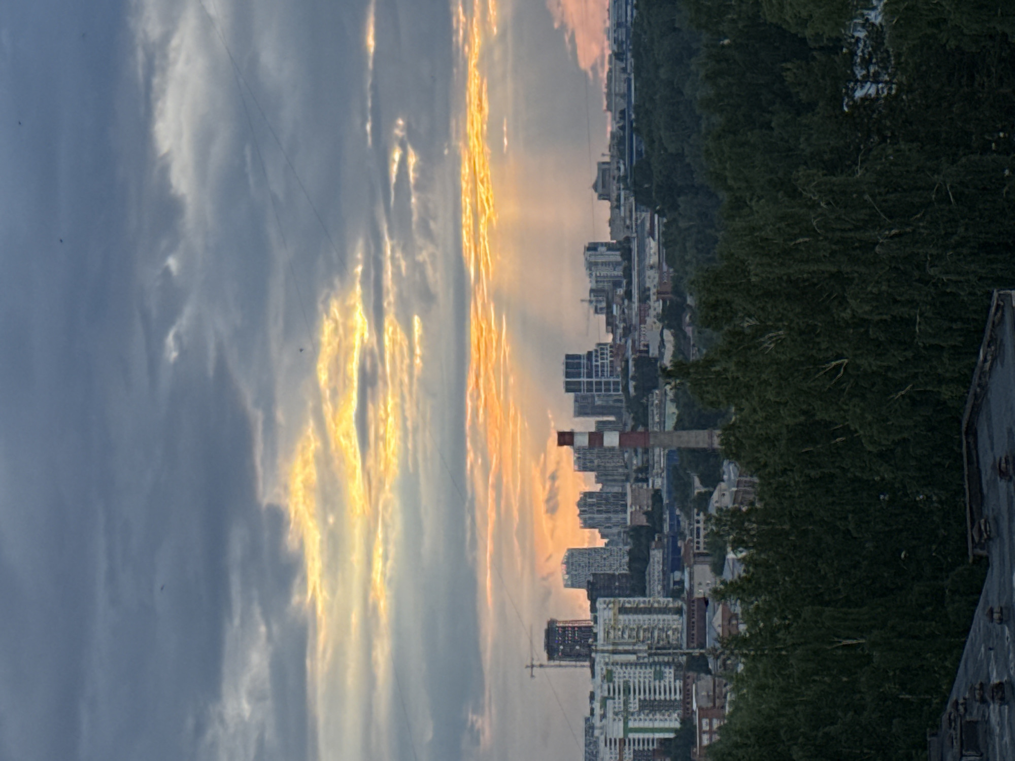 This landscape photograph captures an urban skyline under a dramatic sky at either sunrise or sunset. The sky dominates the upper portion of the frame, displaying a striking contrast between dark, dense grey clouds and brilliant streaks of orange and yellow light from the low sun. The sunlight appears to break through gaps in the clouds, creating visible rays.
Below the horizon, a cityscape stretches across the midground. Numerous buildings, predominantly modern high-rises and apartment blocks, are visible, some silhouetted against the vibrant sky while others are softly lit by the indirect light. A prominent red and white striped industrial chimney stands tall amidst the urban structures, adding a distinct industrial element to the scene. Faint, thin lines, likely power or communication wires, traverse parts of the sky, and a few tiny, dark specks, possibly birds, are scattered within the lighter cloud areas.
On the right side of the frame, a dense, dark green treeline or forested hill rises, partially obscuring the city buildings. In the immediate foreground, on the far right, a dark, angular structure, possibly a rooftop or the edge of a building, is visible. The image conveys a sense of the quiet transition of day to night or vice versa over an urban area. The scene depicts a view of Izhevsk, Russia. No people or specific activities are discernible, and no text is visible.