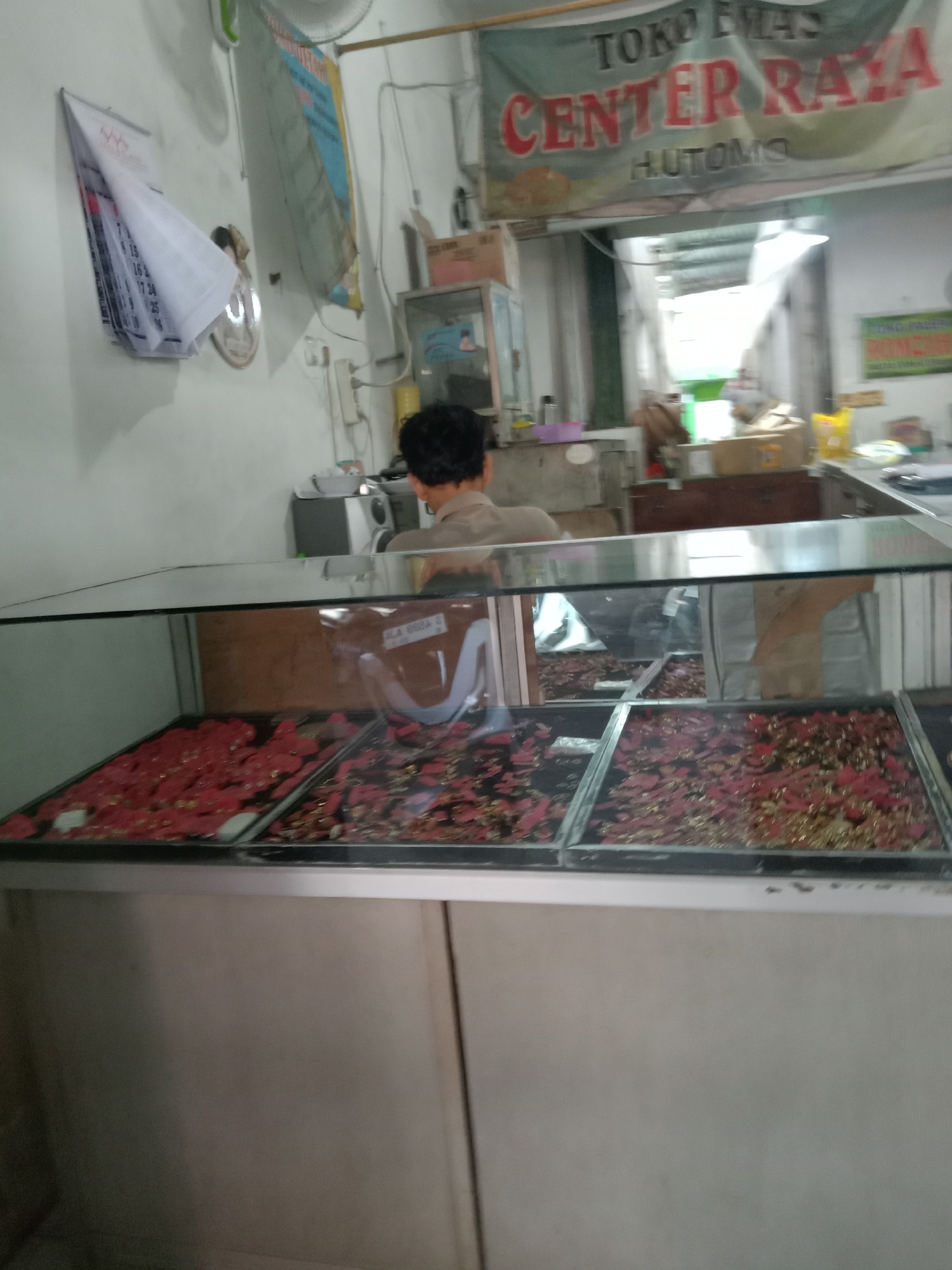 The image depicts the interior of a shop, likely in Sugihwaras, Indonesia, identified by the banner visible in the background that reads "TOKO EMAS CENTER RAYA HUTOMO". A person, seen from behind and wearing a light-colored shirt, is standing behind a glass display counter. The counter contains trays filled with what appear to be various small items, possibly jewelry or decorative beads, with dominant colors of red and gold. In the upper left of the image, a calendar is hanging on the wall. The overall environment appears to be a retail establishment with goods displayed for sale.