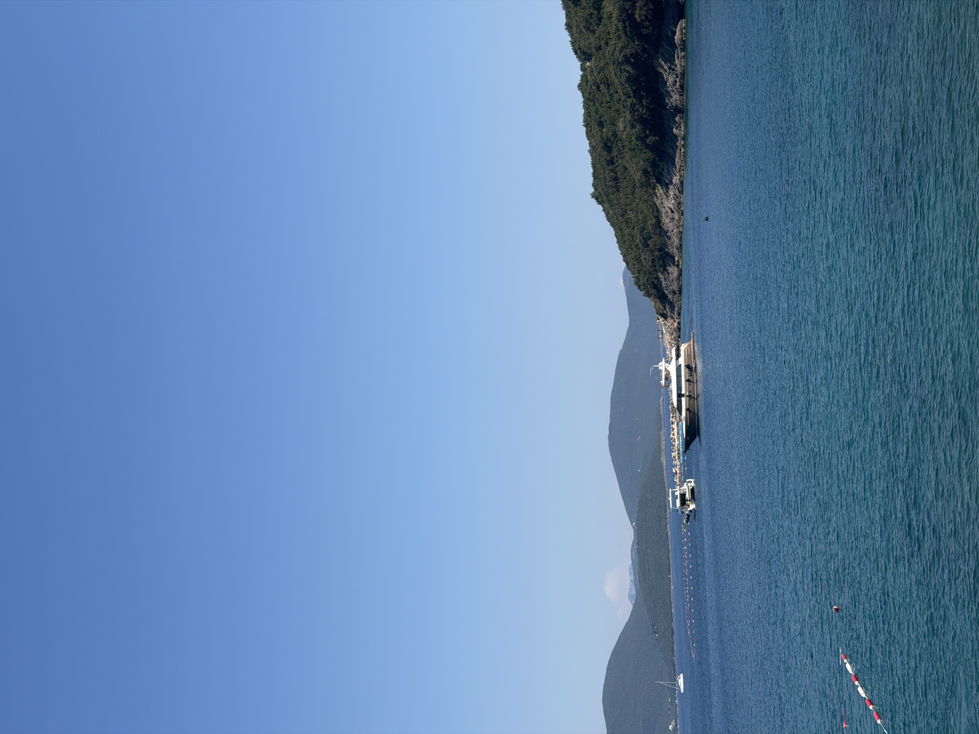 The image depicts a scenic view of the sea near Radovići, Montenegro. The sky is a clear, bright blue, dominating the upper portion of the frame. The sea is a deep, vibrant blue with gentle ripples on the surface, suggesting calm weather. In the middle ground, several boats are visible on the water. To the left, a white sailboat with its sail up is anchored. Further to the right, a white motor yacht with a dark hull is moored, and a larger, dark-hulled motor yacht is positioned further to the right. A series of red and white buoys, potentially marking a swimming area or a channel, are strung across the water. In the background, lush green hills and mountains rise from the coastline, covered in dense vegetation. The shoreline is rocky and appears to be developing, with some structures and breakwaters visible along the coast. The overall impression is one of a tranquil coastal setting on a sunny day, likely during the warmer months.