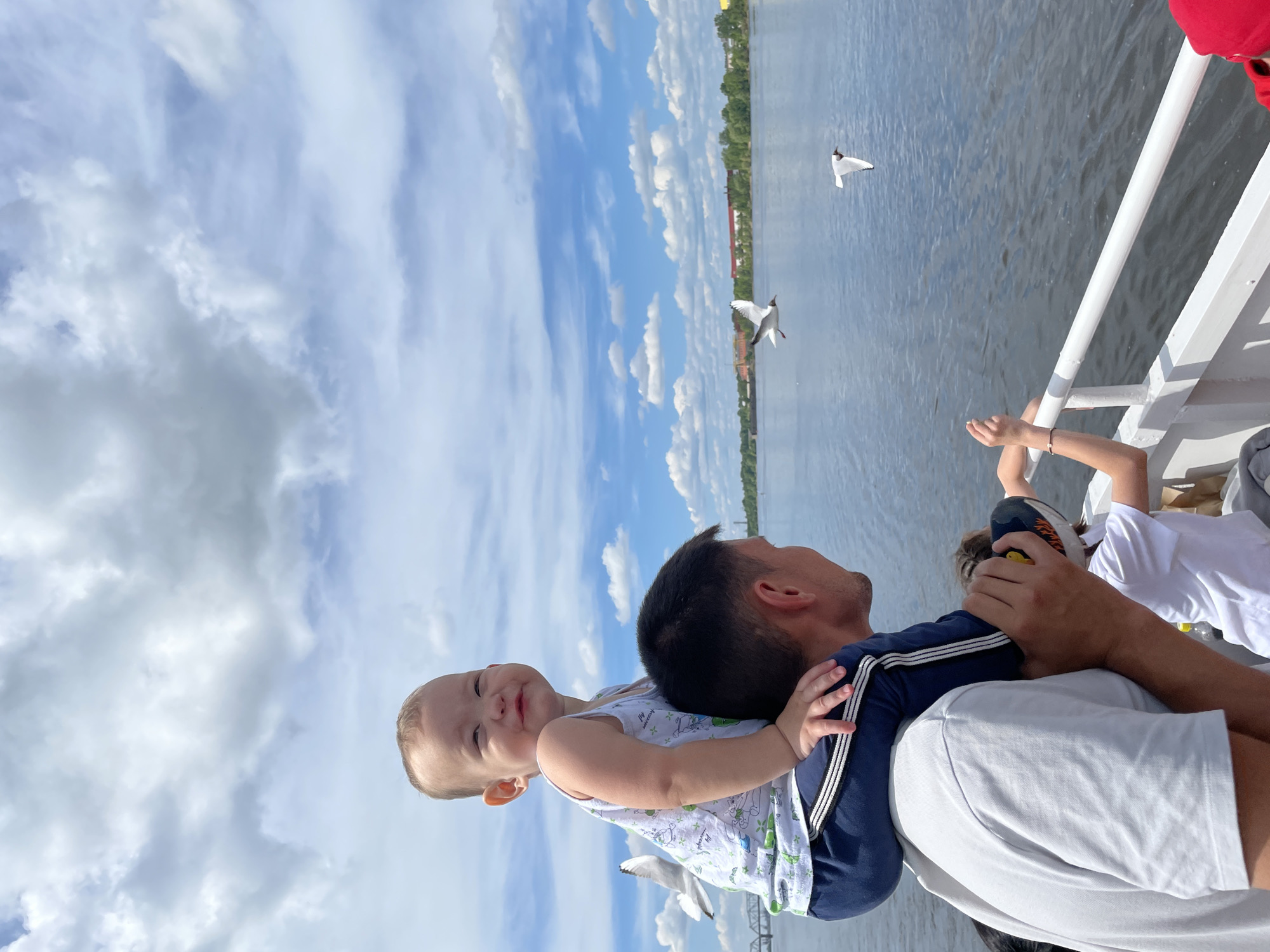 This bright and cheerful image captures a family enjoying a boat ride, likely on the Kama River in Perm, Russia. In the foreground, a man with short dark hair is seen from behind, carrying a baby on his shoulders. The baby, with light hair and wearing a white sleeveless top with light green patterns, is looking directly at the camera with a wide, happy smile, suggesting a joyful moment. The man is wearing a dark blue shirt with white striped details on the shoulders over a lighter t-shirt. To the right, another child is partially visible, wearing a white t-shirt and reaching towards the white railing of the boat.
The setting is a large body of water under a bright blue sky dotted with scattered white clouds, indicating clear and pleasant daytime weather, likely late morning or afternoon. Two white seagulls are visible in flight over the water, adding to the scenic view. Across the water, a distant green shoreline with trees and some buildings can be discerned. The visible white railing suggests they are on a ferry or cruise boat. The overall mood of the image is one of happiness and family bonding during an outing on the water. No visible text is present.