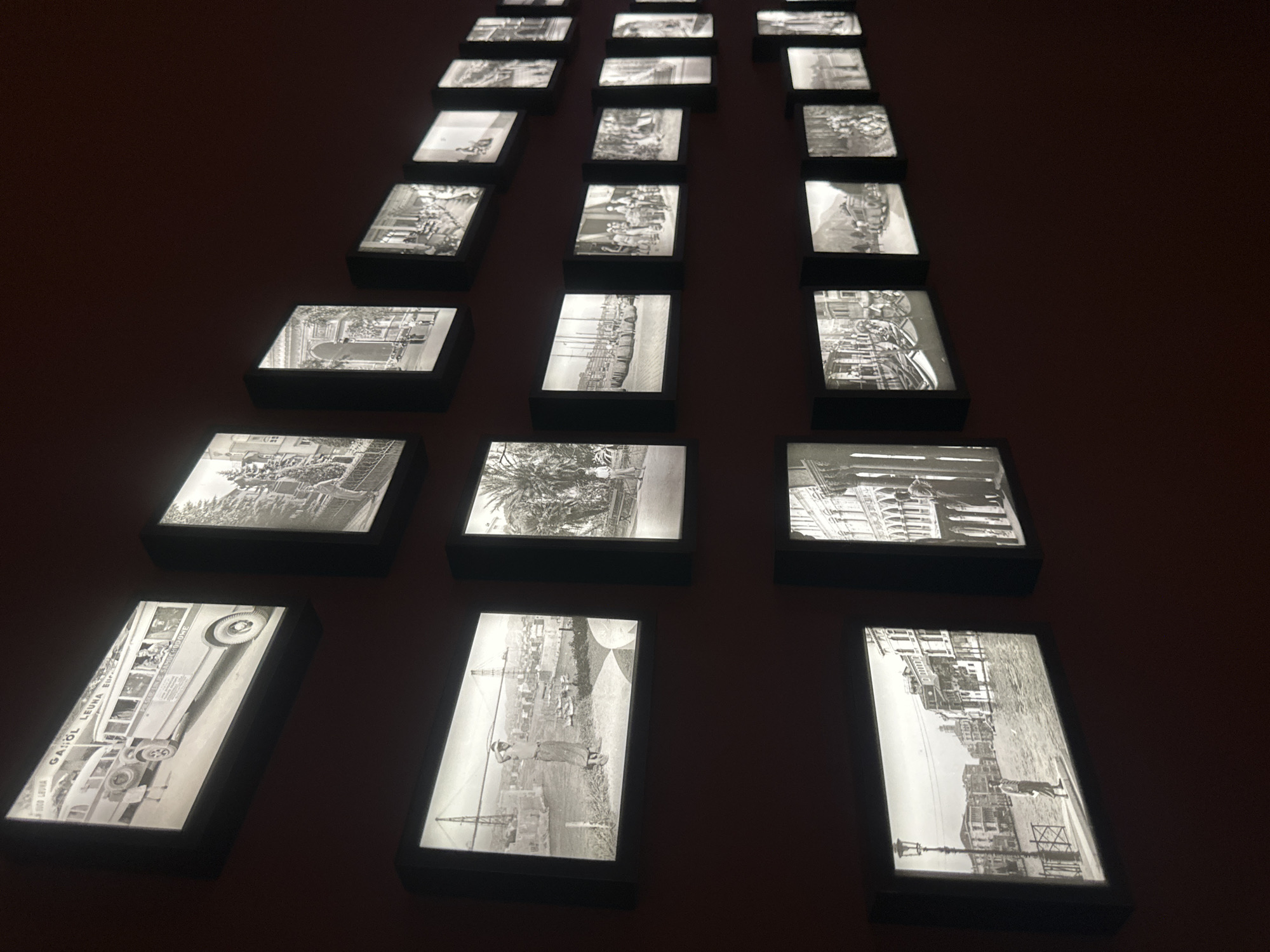 The image displays a gallery of backlit black and white photographs mounted on a dark red wall. The photographs are arranged in a staggered pattern, creating multiple rows. In the top row, the leftmost photograph shows a vintage bus with passengers. The subsequent photographs in this row depict architectural scenes and street views. The second row from the top features images of people and street scenes, including a man standing on a walkway with a suspension bridge in the background. Further to the right, a photograph shows palm trees and two individuals. The third row from the top contains images of boats docked at a harbor, a group of people in an outdoor setting, and architectural elements. The bottom row starts with a photograph of a man standing on a street next to a canal with buildings lining the waterway, reminiscent of Venice. This image is followed by photographs of architectural facades and street scenes. The overall impression is of a curated collection of historical images, likely documenting urban life and historical sites. The reference to Trakai, Lithuania is not visually supported by the provided image.