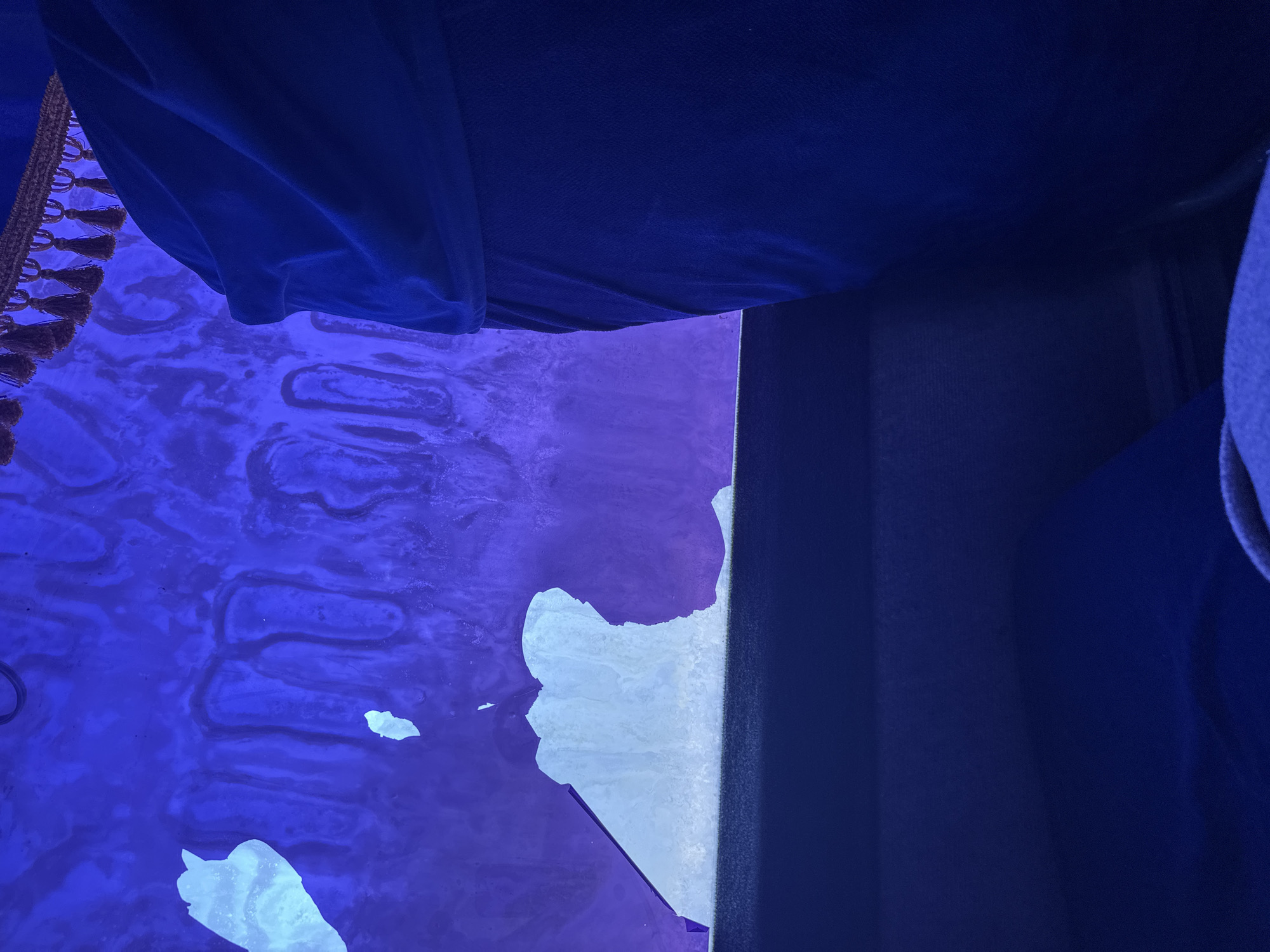 In an indoor setting, illuminated by blue light, a window is visible. The window is partially covered by a light-colored material, possibly frosting or a decal, with irregular patches of transparency. The background beyond the window displays a textured pattern of wavy lines in shades of blue and purple. A dark blue fabric, likely a curtain or upholstery, drapes over the right side of the frame, obscuring a portion of the window and extending downwards. A decorative trim with tassels is visible at the top of the window. The foreground features a dark blue surface, suggesting seating or furniture. The overall scene is dimly lit and abstract, with no people clearly identifiable.