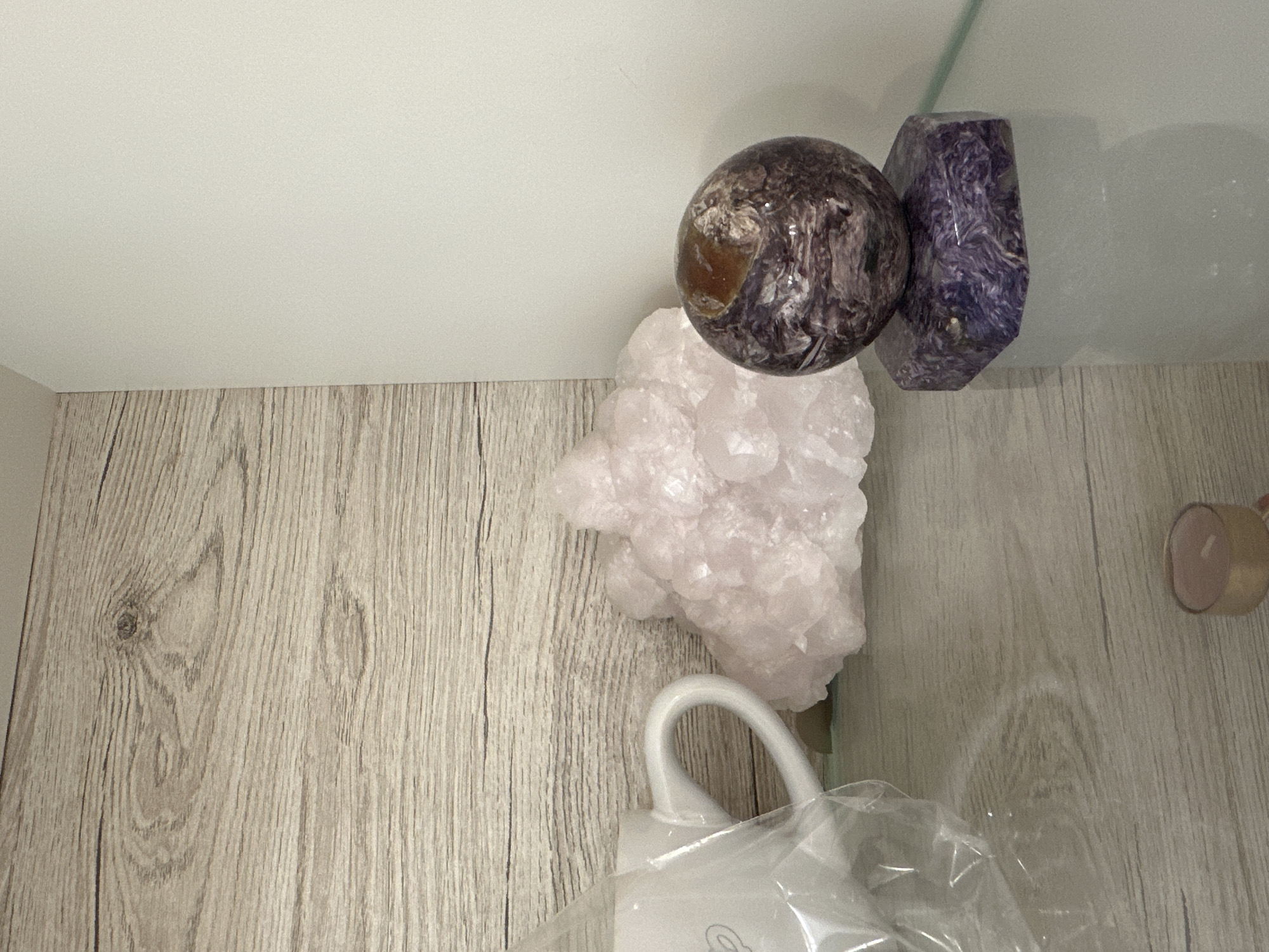 The image shows a corner of a shelf or drawer with a light wood-grain surface. There are three crystals: a sphere with brown and purple swirls, a light pink cluster, and a purple, faceted crystal. There is also a small, gold-colored tealight candle and a white plastic object, possibly a humidifier or diffuser, partially visible in the bottom of the frame and covered in plastic. The location in Sofia, Bulgaria is not determinable from the image.