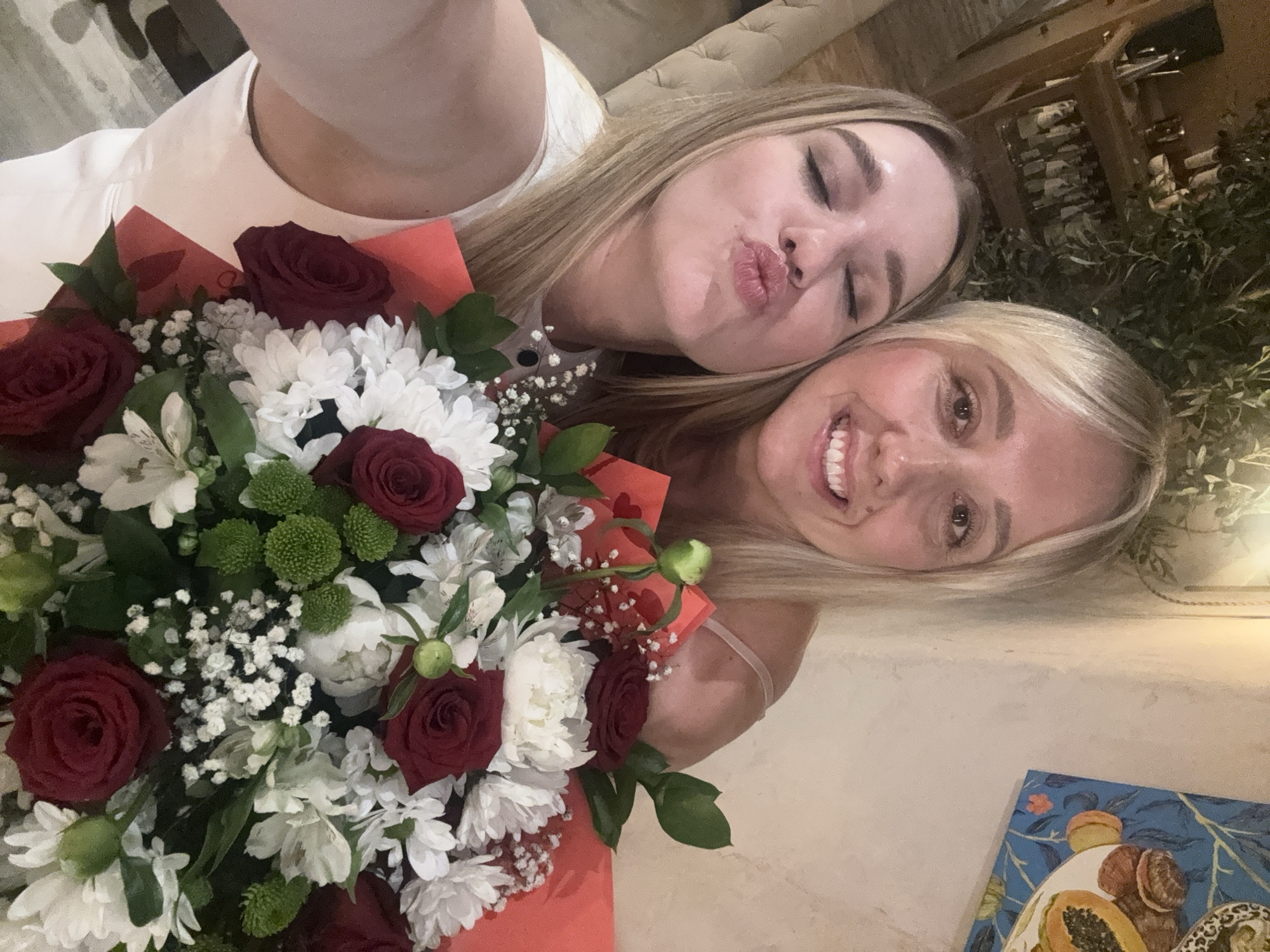 Two women are taking a selfie with a bouquet of flowers. The woman on the left is making a kissing face and has her eyes closed. The woman on the right is smiling broadly. The bouquet is in the foreground and is made up of red roses, white chrysanthemums, and small white and green flowers. The background appears to be an indoor setting with warm lighting, possibly a restaurant or home, with a painting on the wall and some greenery visible. There are no visible elements that suggest the location is in Riga, Latvia.