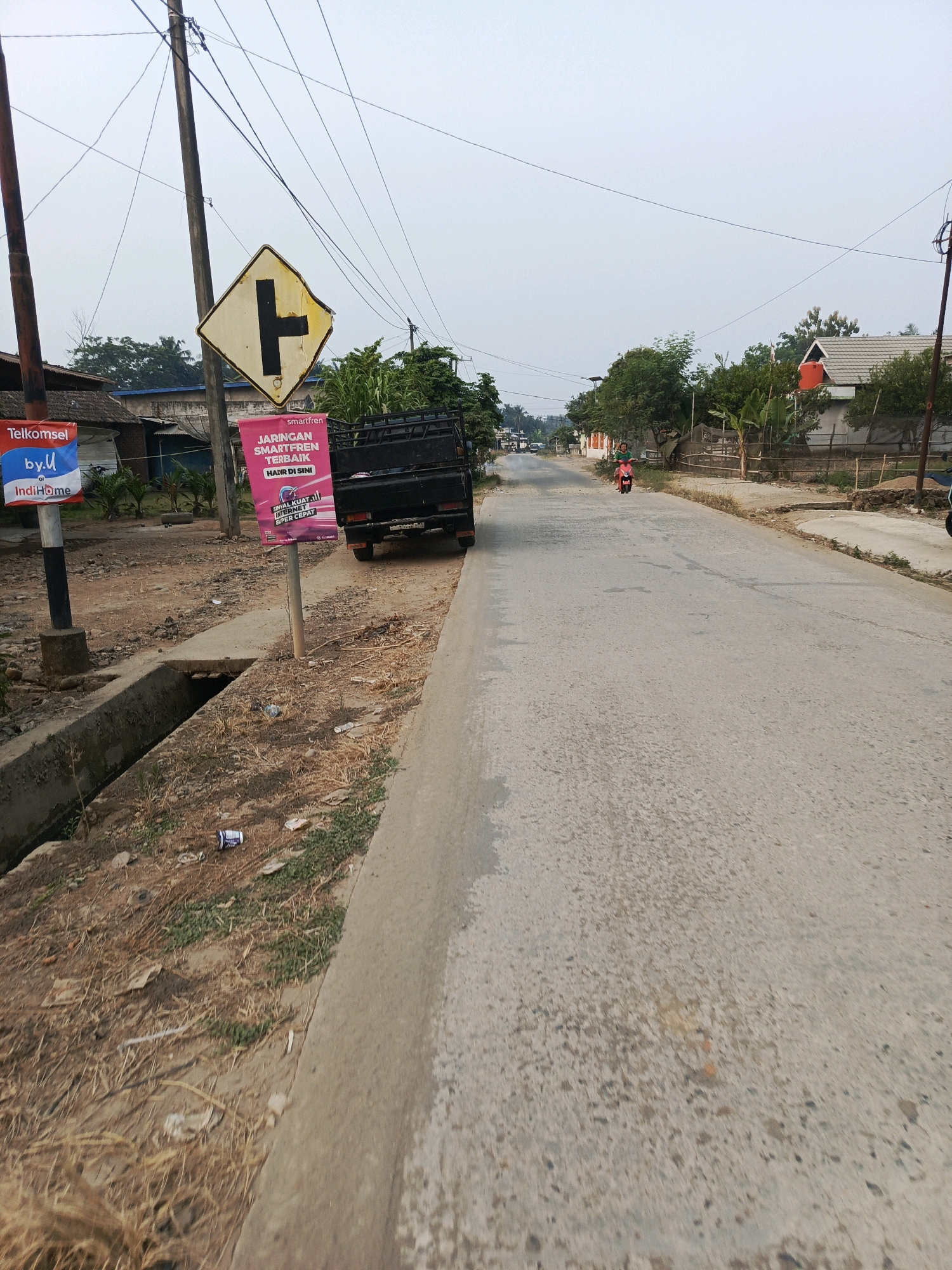 The image shows a roadside scene in Megang Sakti I, Indonesia. The road is paved with concrete and appears to be a two-lane street. On the left side of the road, there is a dirt shoulder with a concrete drainage ditch. Several utility poles with many wires are visible. A yellow diamond-shaped traffic sign with a black "T" intersection symbol is mounted on one of the poles. A black pickup truck is parked on the shoulder. A pink banner advertisement is attached to a pole near the truck. Further down the road, a person is riding a red scooter. Houses and trees are visible in the background. The sky is overcast.