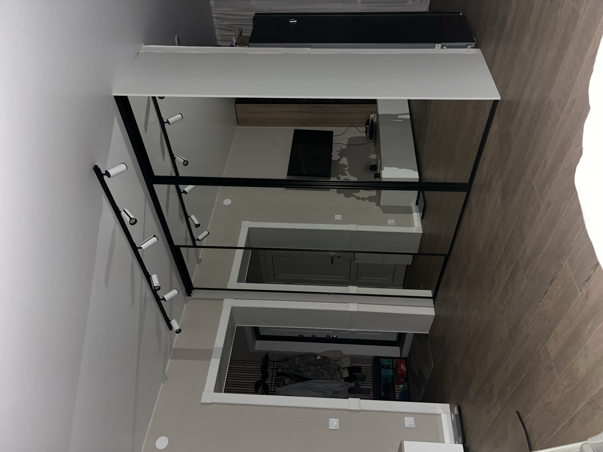 The image shows an indoor scene of a modern living space, possibly an apartment or a house, in an unknown location. The room features a large mirrored wardrobe with black frames that reflects the opposite wall. On the reflected wall, a television is mounted above a white media console. To the left of the wardrobe, there is an entryway with a coat rack holding several jackets and shelves with shoes. The floor is covered with dark brown wooden planks. The ceiling has a track lighting system with multiple adjustable spotlights. The walls are painted in a neutral beige color. The overall impression is one of contemporary interior design with a focus on clean lines and reflective surfaces.