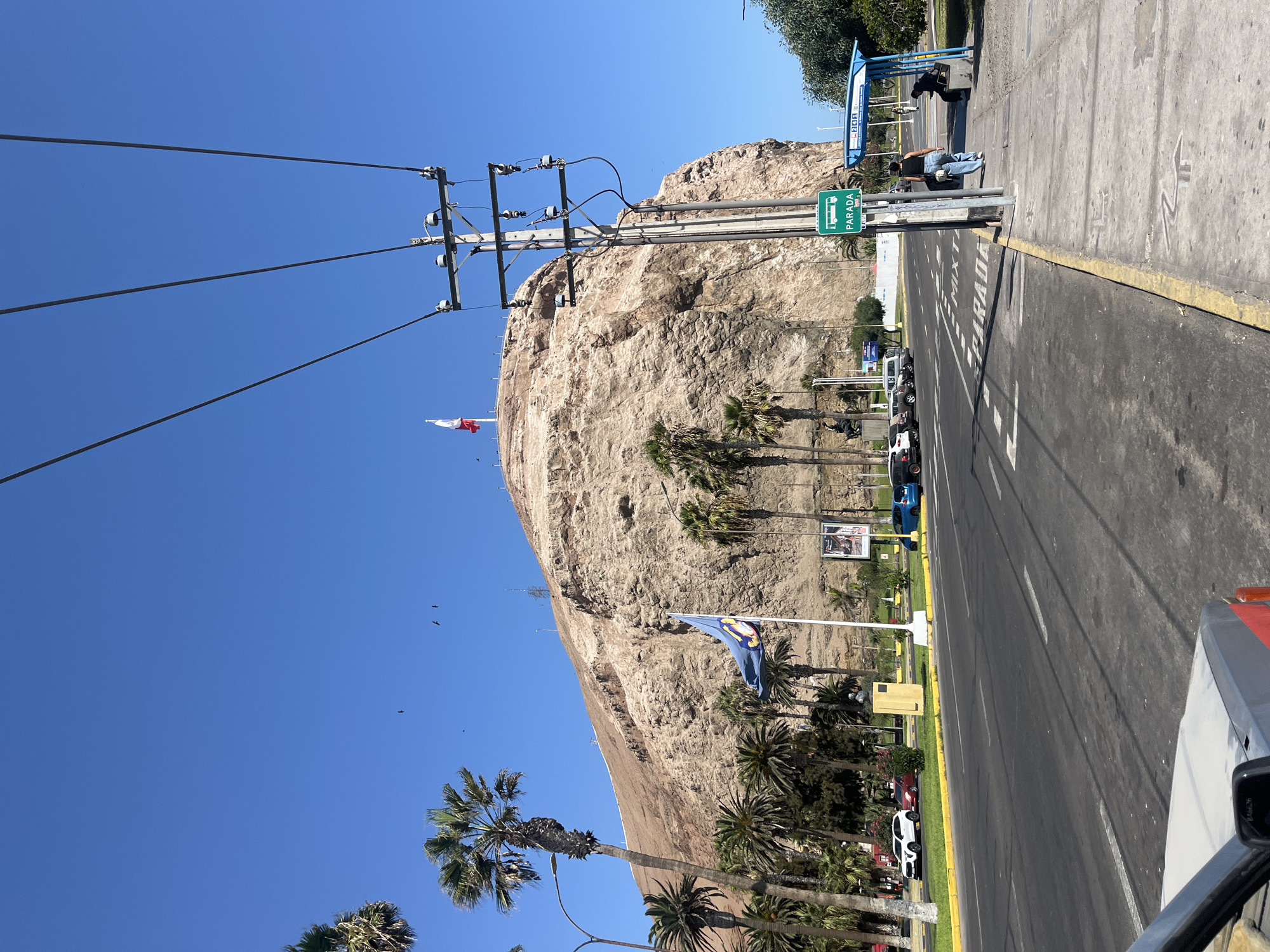 The image depicts a street scene in Arica, Chile, with a large, rocky hill dominating the background. The hill has a Chilean flag at its summit and another flag near its base. Power lines run parallel to the street. Palm trees are prevalent along the side of the road. Cars are parked along the street, and people are at a bus stop. The sky is clear and blue. The image is taken from a slightly elevated perspective, possibly from inside a vehicle. The "Parada" bus stop sign is visible. The overall impression is a sunny day in a coastal city.