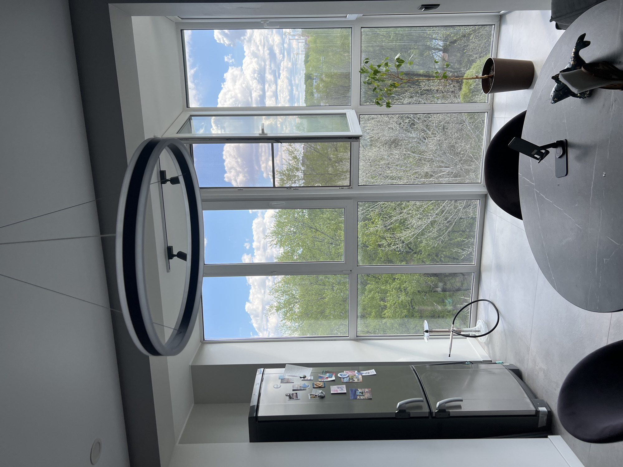The image shows an interior view, possibly of a modern apartment, with a focus on the kitchen area. A large window dominates the background, offering a view of a tree-filled outdoor area under a partly cloudy sky. The window is partially open. In the foreground, a circular modern chandelier hangs from the ceiling. Below the window, a refrigerator with various magnets and pictures on it is visible. Part of a round table with two chairs around it is visible on the right side of the image, and an ornamental plant sits on the table. A glass hookah stands on the floor next to the window. The location is Vologda, Russia based on some of the images on the refrigerator.