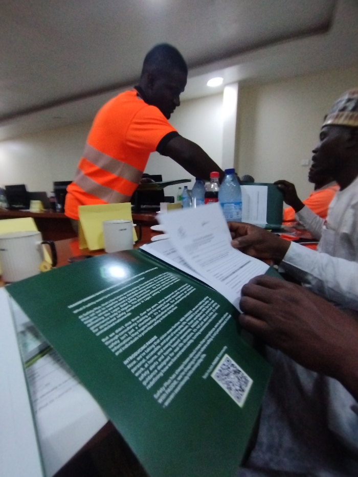 The image shows a meeting taking place at an office in Ashaka, Nigeria. Two men are seated at a table. One is wearing an orange shirt with a reflective vest. The other man, sitting to the right, is wearing a white shirt. The man in the orange shirt is standing up, leaning over the table as if addressing the other man who is seated with a green document in his hands. Multiple water bottles and a Coca-Cola bottle are visible on the table, along with a clipboard, a yellow mug, and a white mug. A few computer monitors are in the background. The image is taken from a low angle, with the focus on the green document.