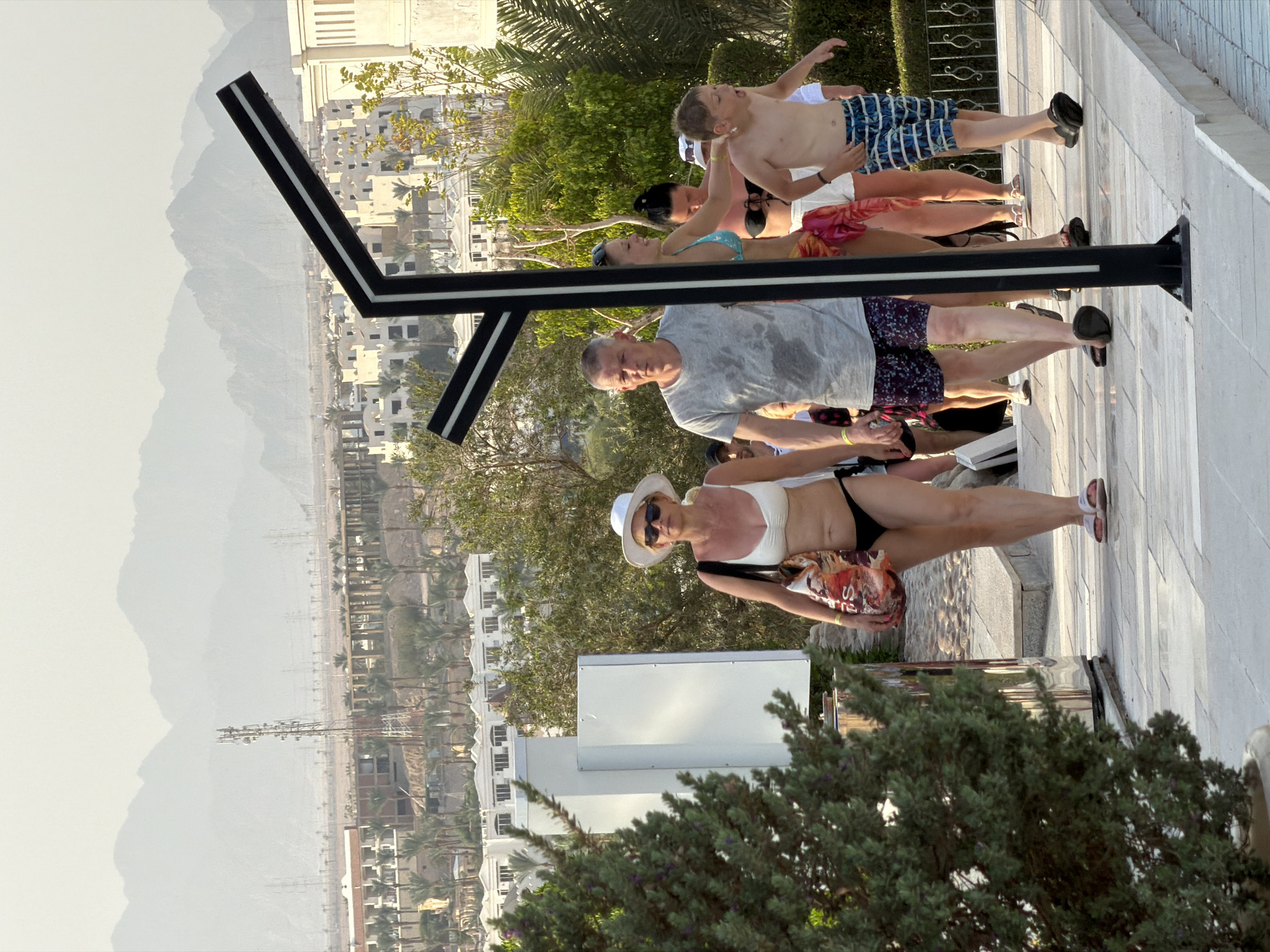 A group of tourists are walking along a paved path in Sharm El Sheikh, Egypt. The path is bordered by a low stone wall and a lush green bush in the foreground. In the background, there are buildings, palm trees, and mountains.
The first person on the left is a woman wearing a white bikini, a white sun hat, and sunglasses. She is carrying a large patterned tote bag. To her right is a man wearing a gray t-shirt and patterned shorts. He has wet patches on his t-shirt.
Behind the man is a woman in a blue bikini and a pink sarong. She is smiling and touching the man's arm. Next to her is another woman in a black bikini.
In front of the man and woman in blue is a young boy wearing swim trunks. He appears to be shouting or laughing. Other people are visible in the background, some walking and some standing.
The overall scene suggests a vacation or resort setting. The presence of palm trees and mountains is characteristic of Sharm El Sheikh, a popular tourist destination in Egypt known for its beaches and diving opportunities. The weather appears to be warm and sunny.