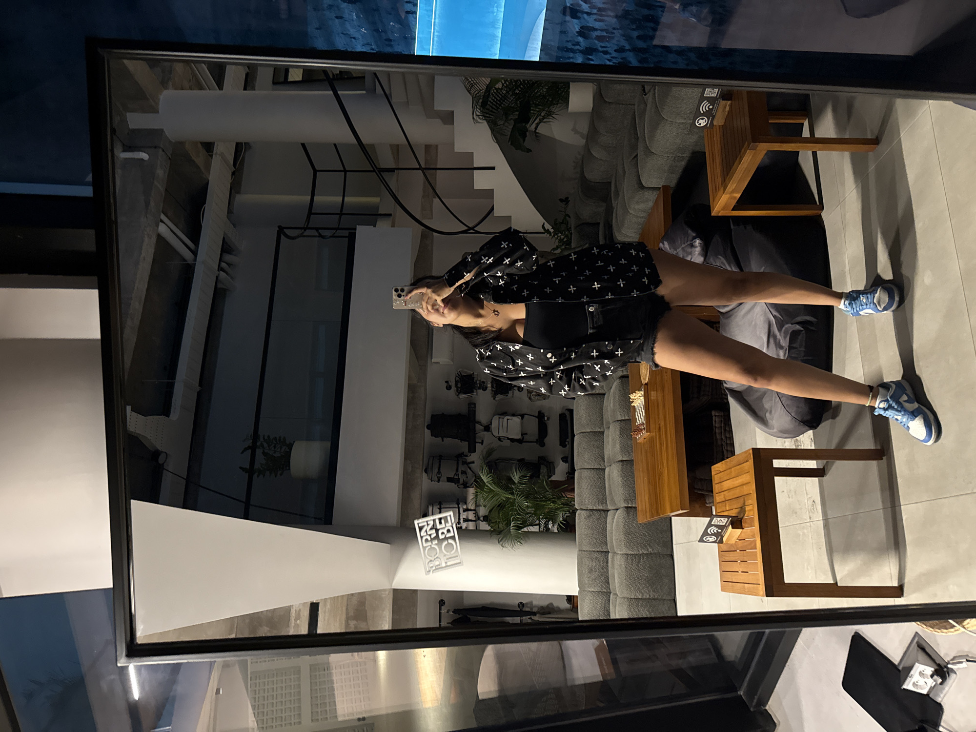 A full-length mirror reflects a young woman in a black patterned jacket, black tank top, and denim shorts. She is taking a selfie with her phone. She is wearing blue and white sneakers. The mirror reflects a modern interior with a grey couch, wooden tables, and a spiral staircase. The location is described as the city of Mengwi, Indonesia, implying the image was taken there. The room appears to be a lounge or waiting area, with plants and a sign that says "BORN TO BE".