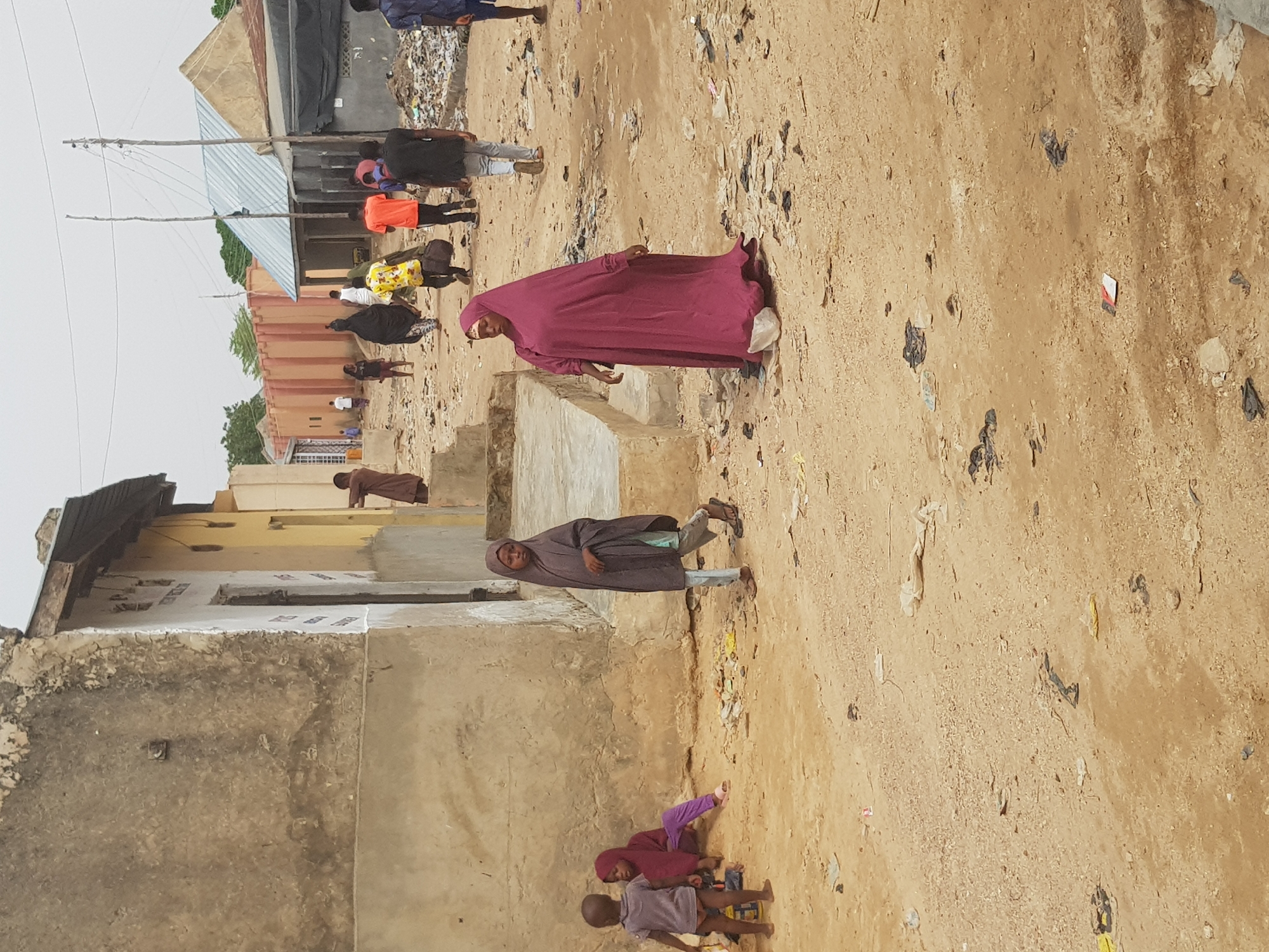 The image depicts a street scene in Dutsinma B, Nigeria. The ground is arid, with scattered trash and debris. Several people are present, including adults and children. One woman in a maroon hijab is walking across a set of concrete steps. Another person in a brown hijab is walking on the ground. There are children playing in the street. Buildings appear in the background, some in disrepair. There are powerlines in the background, and garbage piles.