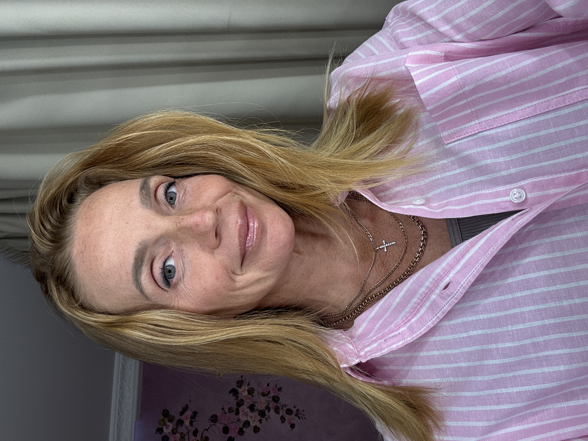 A woman with blonde hair and blue eyes is looking at the camera with a slight smile. She is wearing a pink and white striped shirt and a gold necklace with a cross pendant. The background is blurry but appears to be a room with floral decorations and a curtain. The location is not possible to determine from the image.