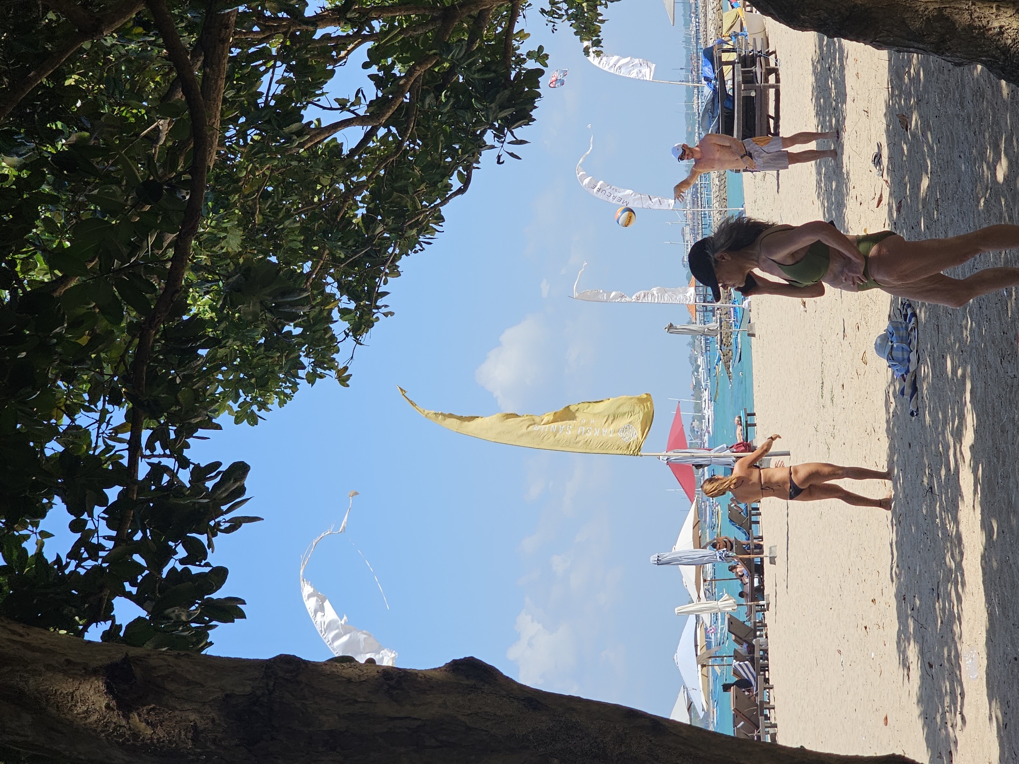 In Denpasar, Indonesia, on a sunny beach, a group of people are enjoying the day. In the foreground, a woman in a green bikini and black baseball cap is on her phone, facing left. Behind her, a man in a white cap and patterned shorts is playing volleyball. Further back, a woman in a black bikini stands on the sand. The beach is lined with lounge chairs and umbrellas. Boats are visible in the turquoise water, and flags flutter in the breeze. The scene is framed by the dappled shade of trees.