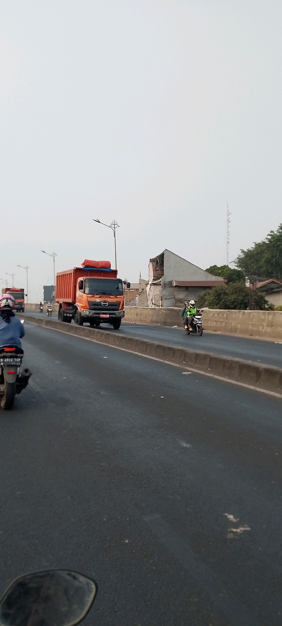 On a bright, overcast day, a multi-lane road is depicted with various vehicles in motion. In the foreground, a motorcycle with license plate B 4972 TXP is visible, with its rider wearing a blue jacket and a helmet. Further down the road, a large orange dump truck with a red tarp covering its load is traveling in the same direction. Behind the dump truck, another orange truck is partially visible. On the right side of the road, a motorcycle carrying two individuals, one wearing a bright green vest, is also moving. The road appears to be elevated, with a concrete barrier separating it from a damaged building with exposed wooden beams on the right. In the background, trees and a distant tower are visible under a pale sky. The scene is consistent with a highway or elevated road in a developed area, possibly Bekasi, Indonesia, given the visible license plate format.