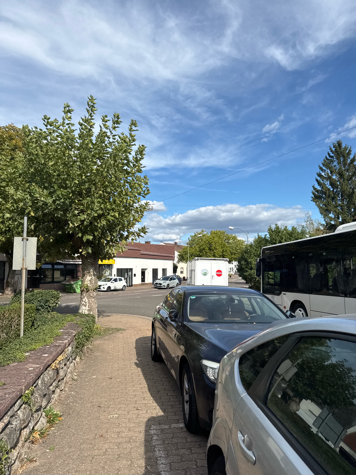 This image depicts a street scene in Überherrn, Germany, on a clear day with scattered clouds. In the foreground, a textured stone wall lines a paved sidewalk. Two cars are parked on the street, a dark sedan partially obscuring a lighter-colored car. Beyond the parked cars, a white bus is visible, its side facing the camera. In the middle ground, a white building with a brown roof has several windows and a doorway. A large tree with green foliage stands prominently on the left side of the image, casting a shadow on the sidewalk. In the background, more trees and buildings are visible under a bright blue sky with wispy white clouds.