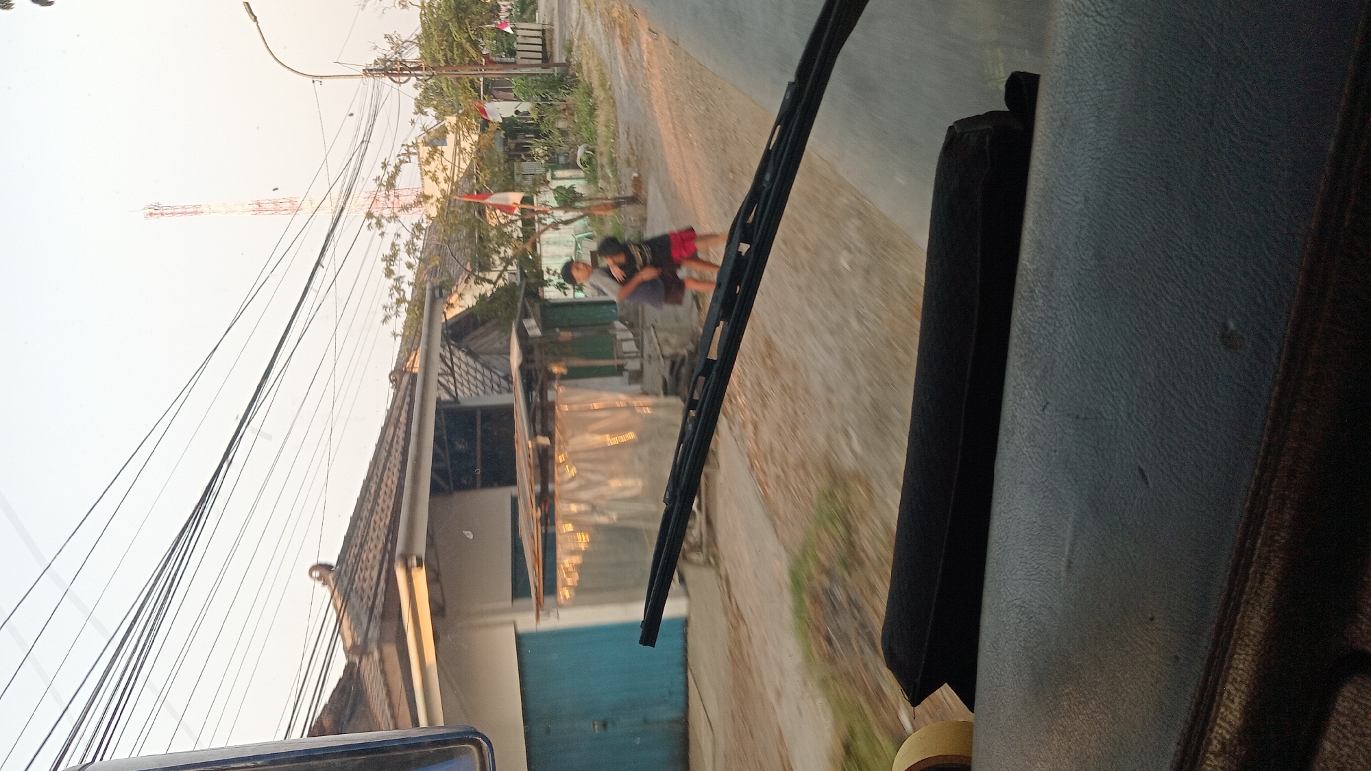 This image captures a scene from within a vehicle, likely a bus or van, looking out through the windshield. The perspective is from the driver's or passenger's seat, with a portion of the vehicle's interior, including a dark upholstered dashboard or seat and a windshield wiper, prominently in the foreground.
Outside the vehicle, the environment appears to be a street in a residential or semi-rural area. Several houses with tiled roofs are visible, one with a light-colored facade and a blue door. A makeshift shelter with a corrugated roof and transparent sheeting is positioned in front of one of the houses. Utility poles and a complex web of overhead wires dominate the upper portion of the image. In the background, a tall telecommunications tower with red and white segments stands against a pale sky. Several Indonesian flags are displayed on poles, suggesting a national holiday or event.
Two young individuals, likely children or teenagers, are the primary human subjects. They are engaged in an embrace or playful tussle on the unpaved ground beside the road. One is wearing a dark t-shirt and shorts, and the other a dark t-shirt and red shorts. The lighting suggests it is either early morning or late afternoon, with soft, warm sunlight casting shadows. The unpaved road and the general state of the buildings suggest a location in a developing area, consistent with a place like Jagong, Indonesia, which is known for its rural and developing characteristics.