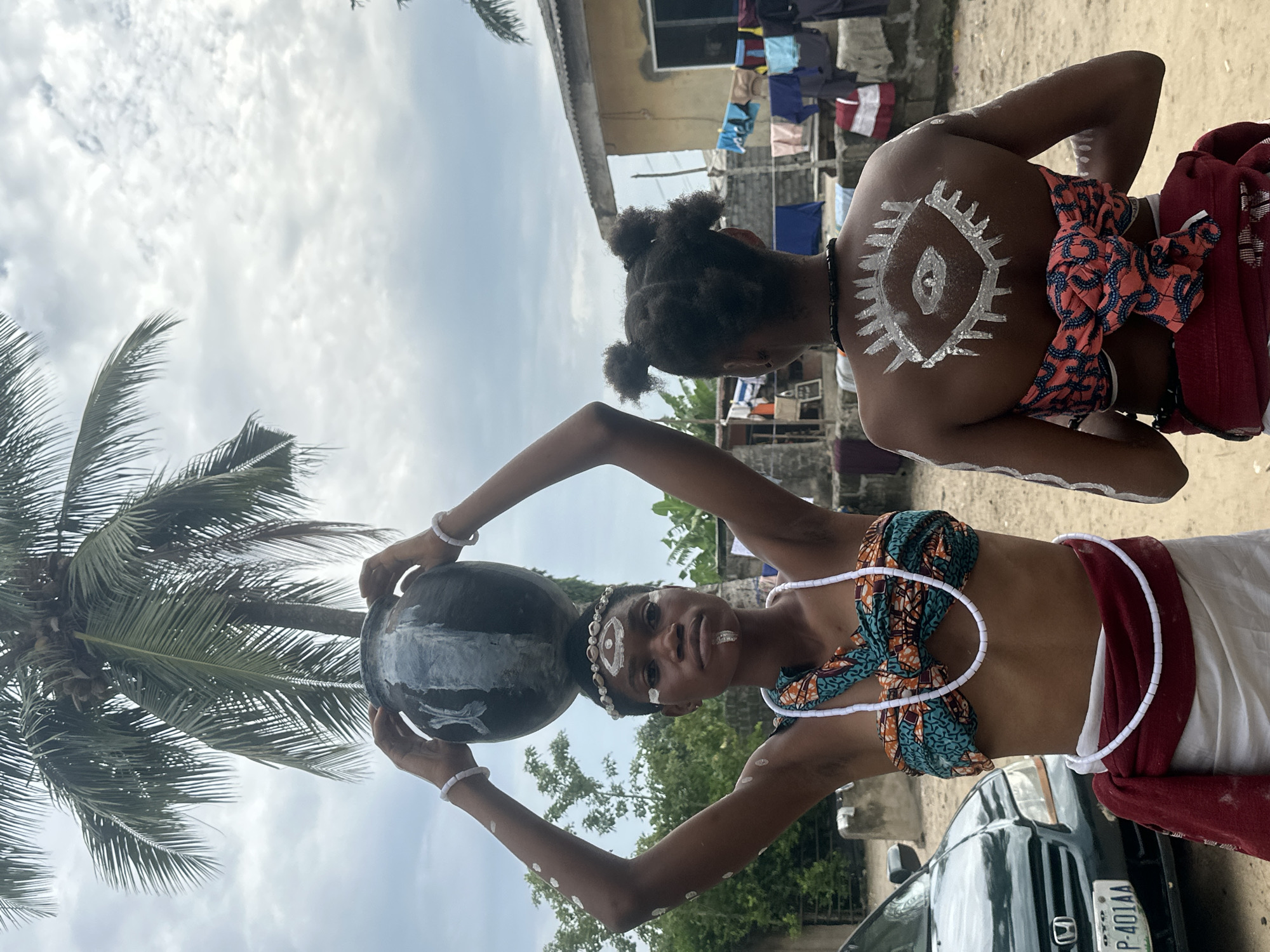 Two young women are depicted from the back and side, adorned with white body paint in symbolic designs, suggesting a cultural or ritualistic context. The woman on the left balances a large, dark pot on her head, her arms raised to support it. She wears a colorful, patterned top with a white beaded necklace and a red sash tied around her waist. The woman on the right has a stylized eye painted on her back, with smaller painted markings on her arms. She wears a similar patterned cloth tied around her waist and a dark beaded necklace.
The setting appears to be an outdoor area in Badagry, Nigeria, characterized by a sandy ground, a prominent palm tree, and a building with laundry hanging on a line. A dark green car is parked to the left of the frame. The sky is overcast with clouds. The overall scene conveys a sense of cultural tradition and possibly a performance or ceremony taking place in a residential or communal outdoor space.