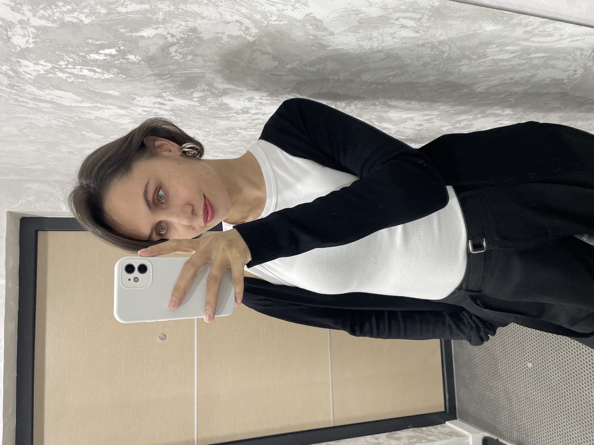 A woman is taking a selfie in what appears to be a bathroom or powder room. She is holding a white smartphone in her left hand, positioned in front of her to capture the image. She is wearing a white tank top, a black cardigan, and dark pants with a visible belt buckle. She has short brown hair and wears hoop earrings. The background includes a bathroom mirror with a black frame, and the wall behind her is textured in a light gray and white pattern. The bottom right of the frame shows a grid-like texture, which may be a part of a bathroom fixture or ventilation system. The location is not discernible as Izhevsk, Russia, based on the visual information provided in the image.
