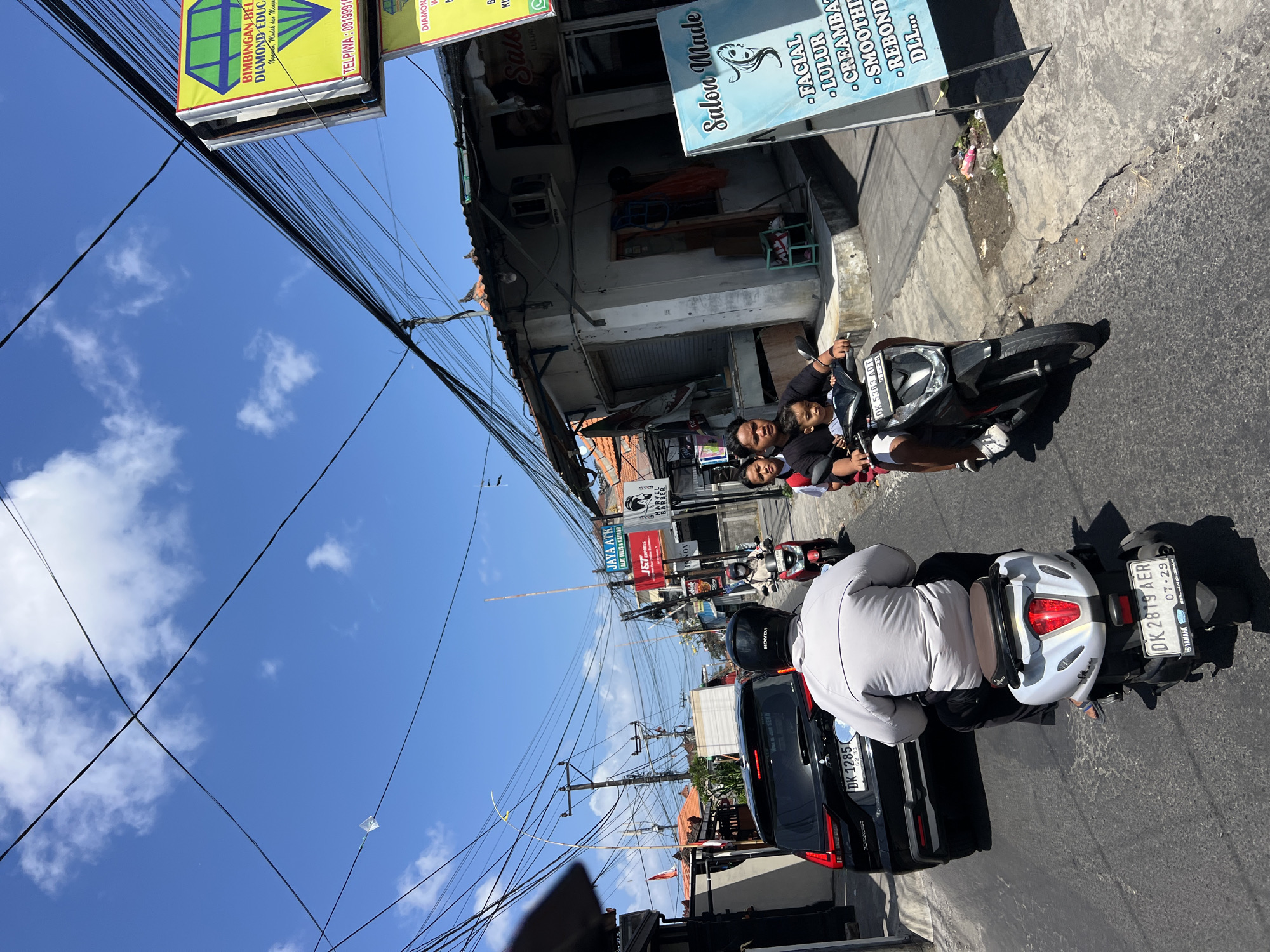 In Denpasar, Indonesia, on a sunny day with scattered clouds, a busy street scene unfolds. Several vehicles, including cars and scooters, navigate the asphalt road. One scooter carries a family of four: a man driving, a woman behind him, and two children in front of the woman. Another scooter follows, carrying a man wearing a helmet and a grey jacket. A black SUV is parked on the side of the road, with its license plate clearly visible as DK 1285. Overhead, a complex network of power lines crisscrosses the sky.
Along the street, buildings house various businesses. A sign for "JAYA ATR" and "J&T Express" is visible, as is a barber shop sign for "HARVAN BARBER". Further down, a brightly colored sign advertises "BIMBINGAN BELAJAR DIAMOND EDUCATION". Another sign, a light blue one with white text and an illustration of a woman's profile, promotes "Salon Made" and lists services such as "FACIAL", "LULUR", "CREAMBATH", "SMOOTHING", "REBONDING", and "DLL...". The storefronts suggest a commercial area with a mix of services and educational facilities. The overall impression is of a vibrant, everyday street in a bustling Indonesian city.