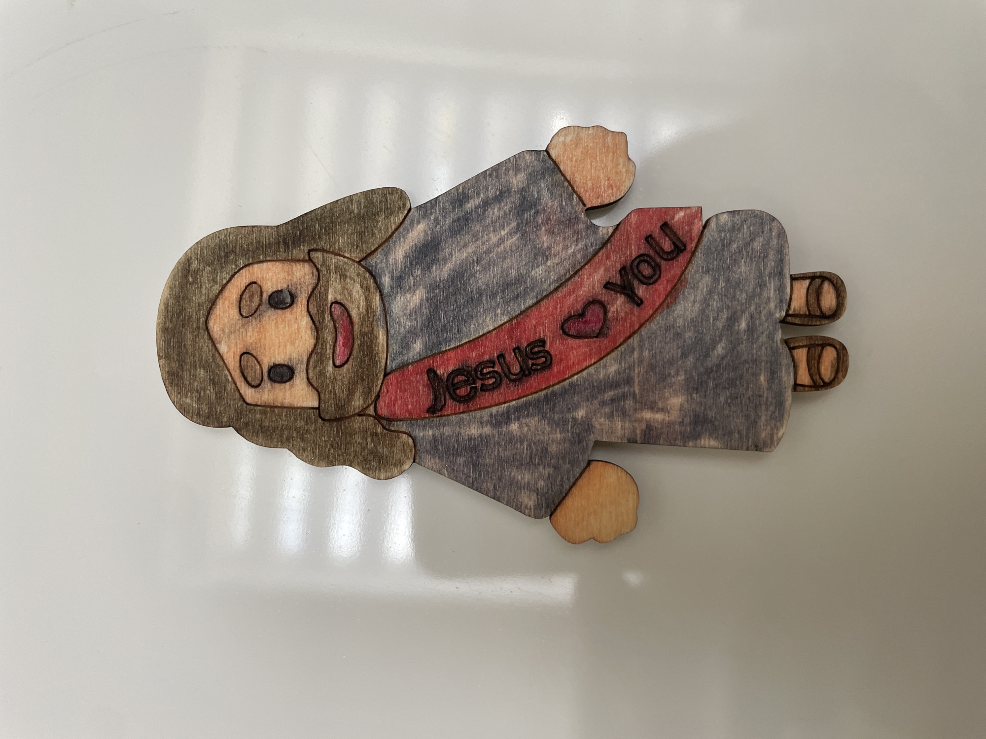 The image shows a wooden cutout of Jesus Christ, depicted as a cartoonish figure with brown hair and beard, a grey robe, and brown sandals. A red sash with the words "Jesus loves you" and a heart symbol is draped across his chest. The cutout is placed on a white, reflective surface, likely a table or countertop. There are no visible contextual clues to indicate its location in the city of Cahul, Moldova.
