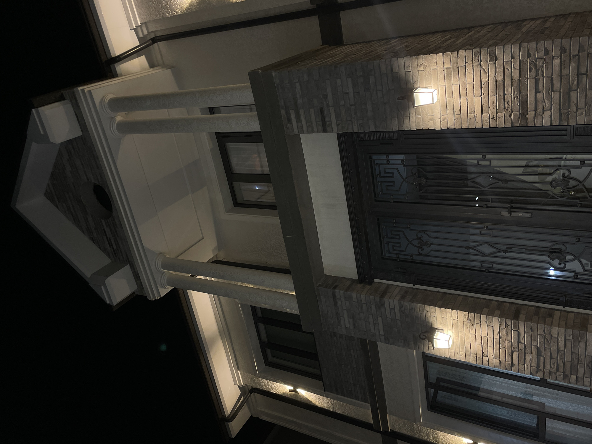 The image shows the facade of a modern house at night. The house features a prominent entrance with a large double door with decorative metalwork. Flanking the door are brick pillars illuminated by warm-toned wall sconces. Above the entrance, there is a balcony supported by two large columns and a pediment. Windows are visible on the upper level, reflecting the exterior lighting. The overall architectural style appears to be a blend of classical and contemporary elements. The specific location in Tashkent, Uzbekistan cannot be determined from the image alone.
