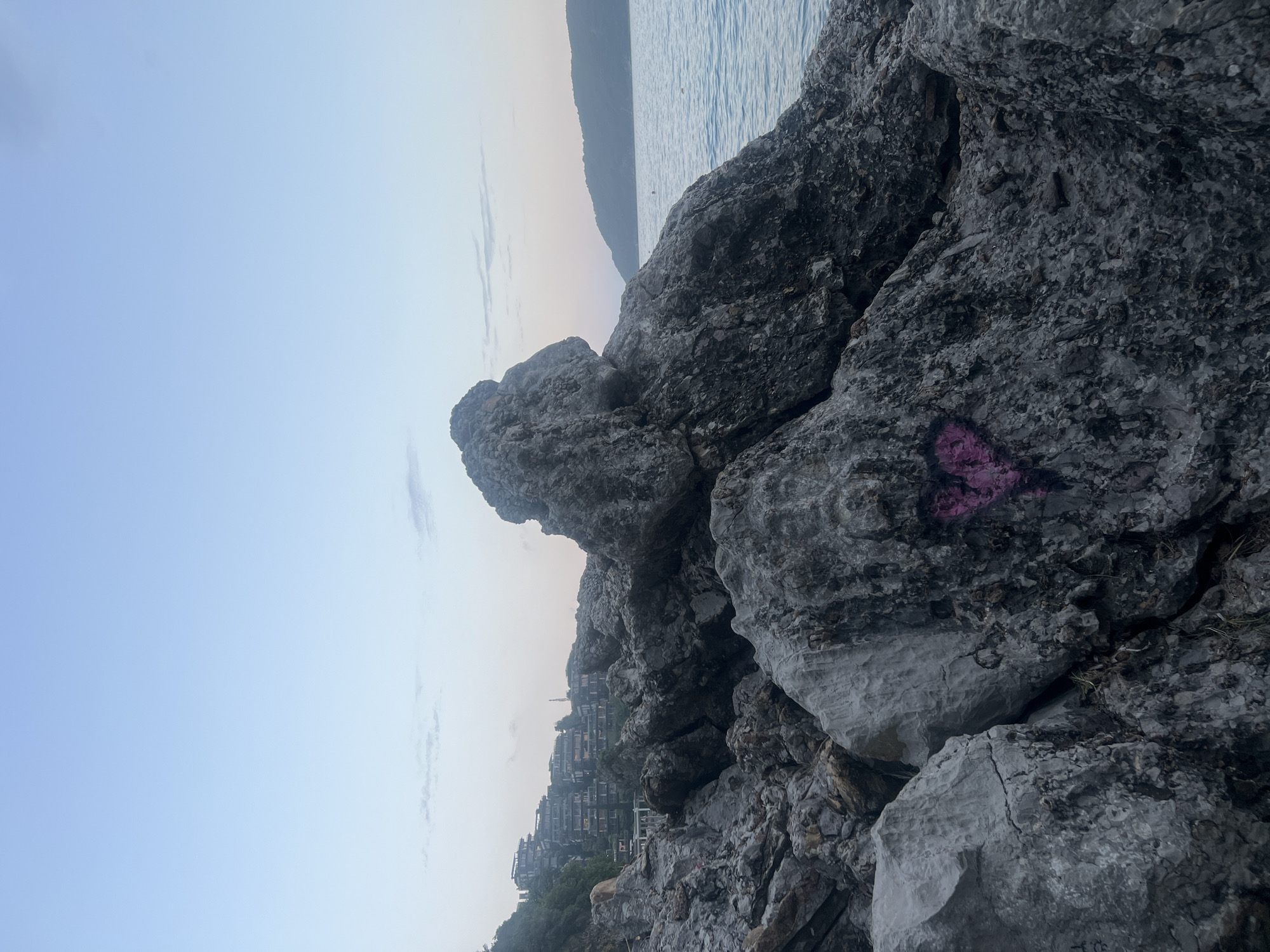 The photograph presents a view in Budva, Montenegro, featuring a rocky shoreline along the Adriatic Sea. Large grey rocks dominate the foreground, some marked with graffiti including a pink heart. In the middle ground, a hillside is densely built with residential structures. The background reveals the sea extending towards a mountainous landmass under a hazy sky. The lighting suggests either dawn or dusk. No people are visible in the frame.