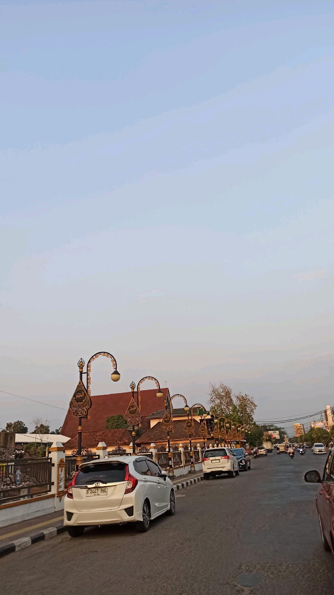 The image shows a street scene in Surodikraman, Indonesia, during the late afternoon or early evening. The sky is a clear, pale blue. On the left side of the street, a white Honda Jazz is parked. Behind it, a low wall with decorative metalwork lines the property, and a building with a reddish-brown tiled roof and ornate streetlights with Islamic calligraphy designs is visible. Several other cars are parked or driving on the right side of the road, which is paved and has a white and black striped curb. Further down the street, a traffic light and buildings can be seen. Motorcycles are also present in the traffic. The overall impression is of a typical urban streetscape in Indonesia.