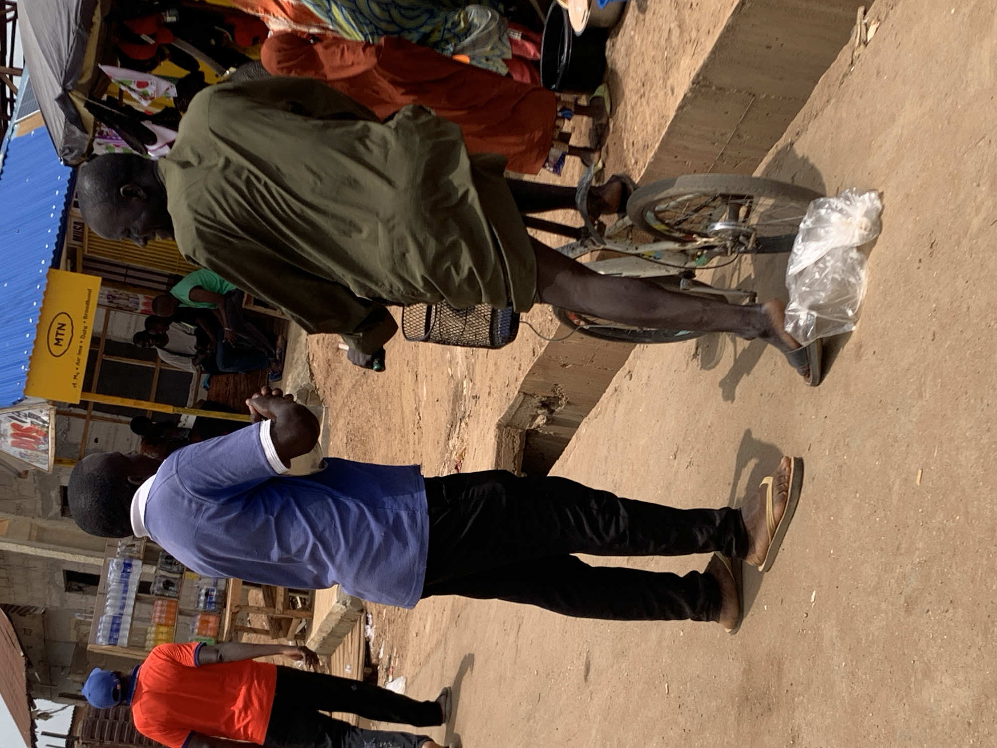 In Akwanga, Nigeria, a man stands in a blue shirt and black pants in front of an MTN shop and water vendor. A second man on a bicycle is riding away, barefoot, with a bag on the front of the bike. An orange-shirted man walks by. The ground appears to be dirt or a concrete-like surface.
