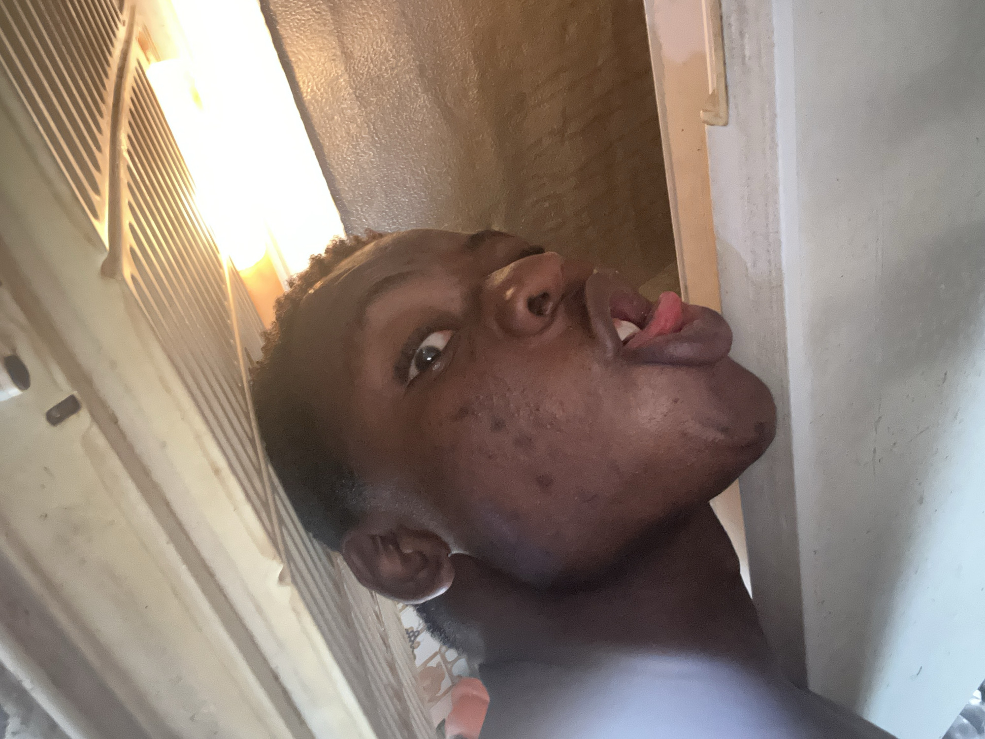 A medium close-up portrait shows a dark-skinned person making a face with their tongue sticking out. The person's head is tilted upwards and angled to the right. The background consists of a wall with a door frame and what appears to be a textured surface. The location is not identifiable.
