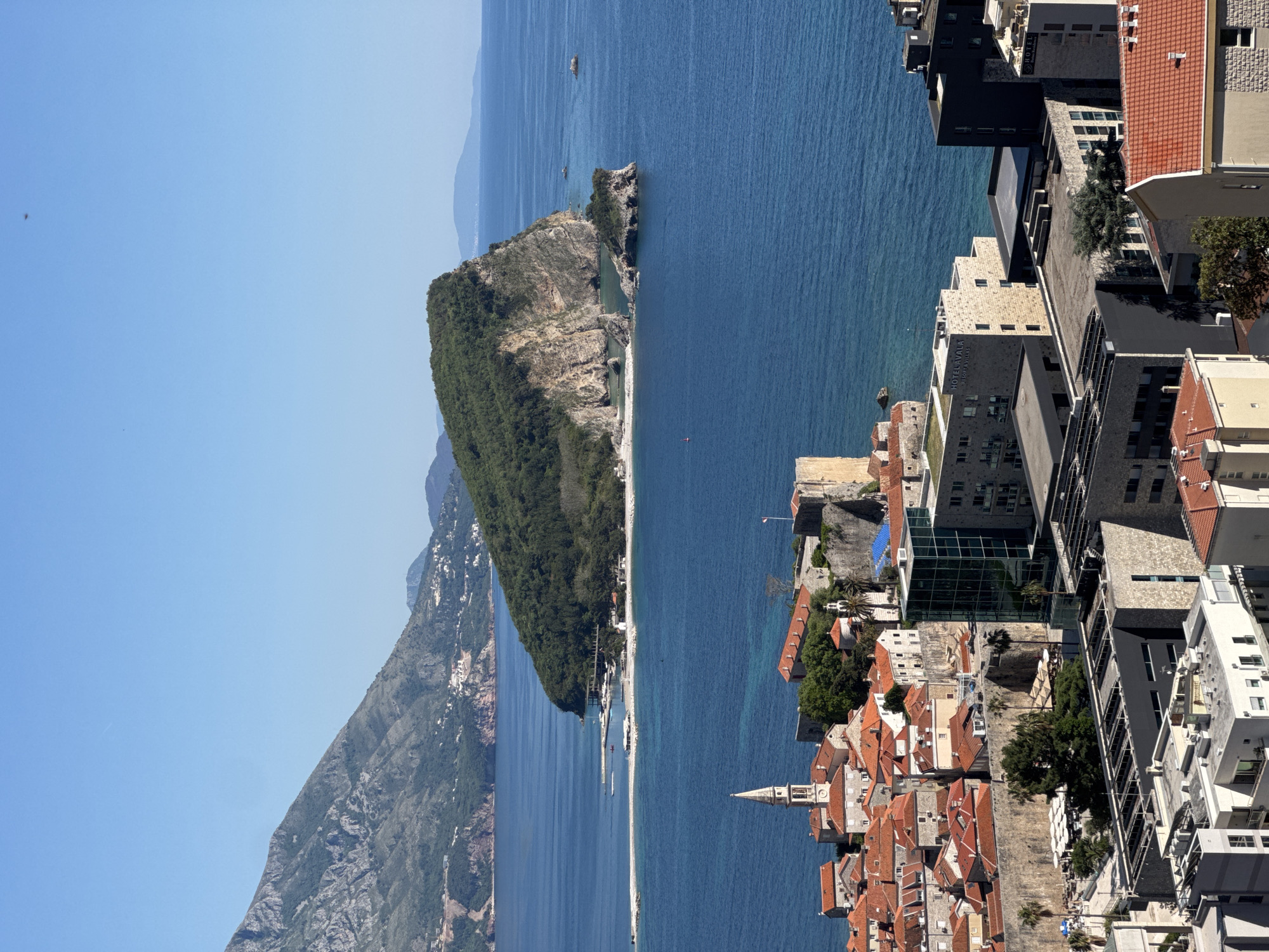 The image shows an elevated view of the coastline in Budva, Montenegro. The Adriatic Sea extends into the distance under a clear blue sky. To the left, a peninsula covered in dense green forest stretches out into the sea. A narrow strip of beach lines the base of the peninsula, with small boats docked offshore.
On the right side of the image are the buildings of Budva, characterized by red-tiled roofs, stone facades, and modern architectural elements. The architecture reflects a blend of historical and contemporary styles. A tall spire or tower rises above the rooftops near the shore.
The picture captures the contrast between the natural landscape of the peninsula and the urban development of the city, with the blue sea forming a central connecting element.