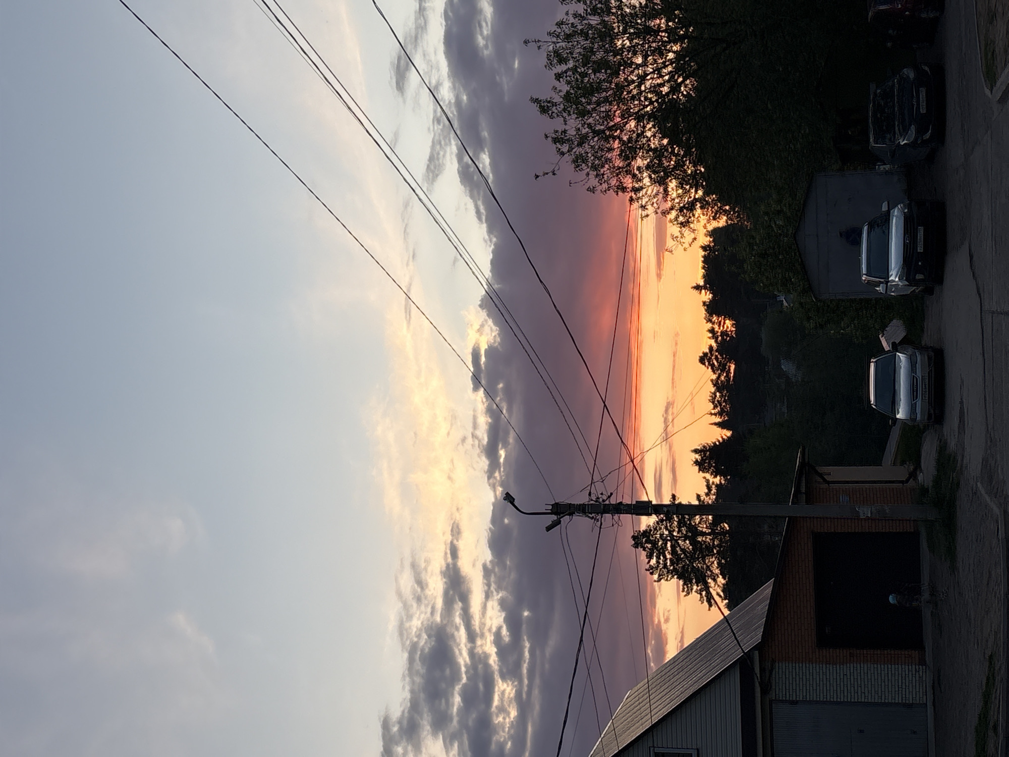 The image shows a street scene in what appears to be a residential area in the city of Chaikovsky, Russia. A sunset is in the background, with hues of orange and pink visible through the clouds. Power lines crisscross the sky, attached to a utility pole located near a brick and siding building with a garage door. Three parked cars are visible further down the street. Trees partially obscure the view of some of the buildings and the cars. The image has been rotated.