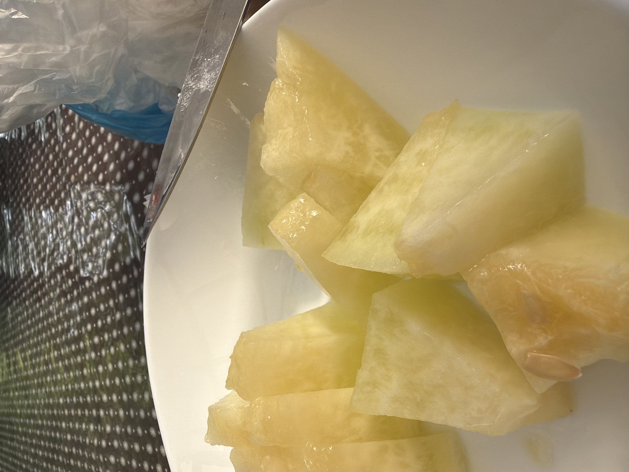 A close-up shot captures several chunks of peeled yellow fruit, likely honeydew melon, arranged on a white plate. The fruit is sliced into irregular, somewhat angular pieces, some revealing a slightly translucent interior. A sliver of a metal knife is visible at the top right corner of the plate, suggesting the fruit has recently been cut. In the background, a patterned surface, possibly a tablecloth or placemat with a dot pattern, is visible, along with a blue plastic bag and white plastic wrapping. The lighting is soft, highlighting the juicy texture of the fruit. The specific location in Sochi, Russia is not discernible from the image itself.