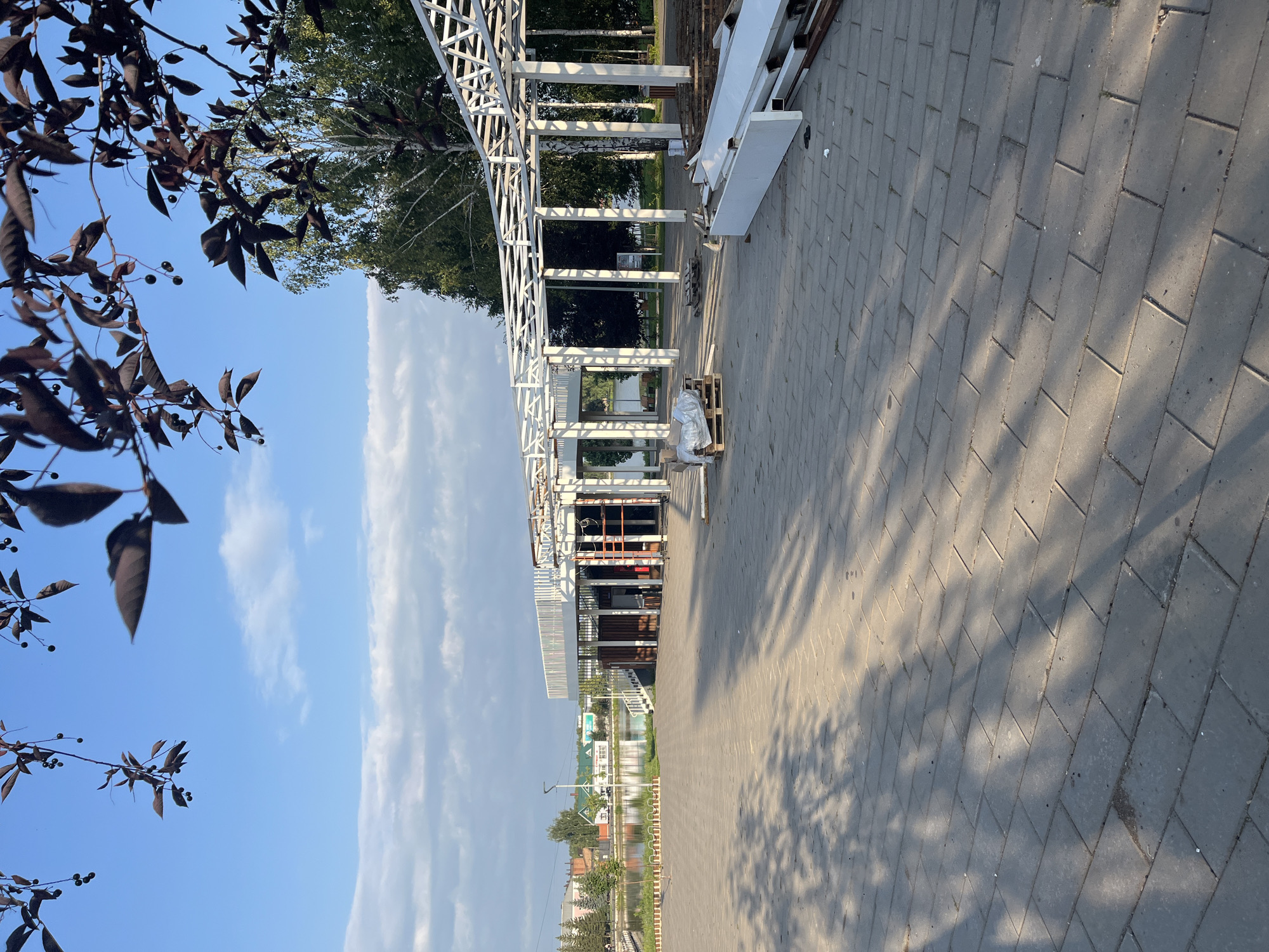 The image depicts an outdoor scene in Novye Turbasly, Russia, featuring a paved area with a large, partially constructed white metal structure resembling a pergola or canopy. The structure has a modern, open design with multiple support columns and intricate geometric crossbeams. In the foreground, the paved ground is covered in gray rectangular tiles, with dappled shadows cast by nearby trees. Construction materials, including white metal beams and wooden pallets, are scattered on the ground near the structure, indicating ongoing work. To the left, a body of water, likely a lake or river, is visible, with trees and buildings lining its far bank. The sky is blue with scattered white clouds, and the branches of a tree with dark leaves and small berries hang into the frame from the top. The overall impression is one of a public park or recreational area undergoing development. There are no people visible in the image.