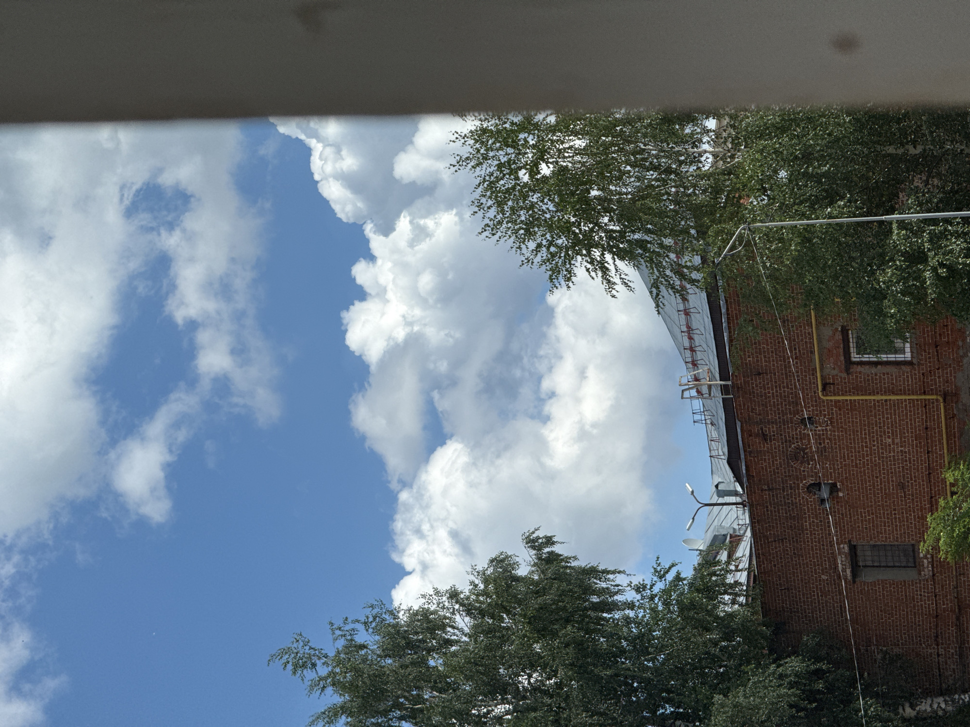 This is a daytime, medium-angle shot taken from an elevated perspective, looking up at a brick building with trees and a bright blue sky with fluffy white clouds. The building is made of red brick and appears to be older. On the roof of the building, there are what appear to be industrial structures, including metal roofing, pipes, and a streetlamp. Two large trees, likely birch trees given their distinctive bark, flank the building. The tree on the left is closer to the viewer and larger, its branches reaching up towards the sky. The tree on the right is smaller and further back. The sky is a vibrant blue, dotted with scattered white cumulus clouds. A light-colored, vertical pillar is visible in the right foreground, partially obscuring the view of the sky and the right side of the building. The scene is static and appears to capture a moment of calm on a clear day. The image does not contain any people or discernible actions. The location is identified as the city of Votkinsk, Russia.