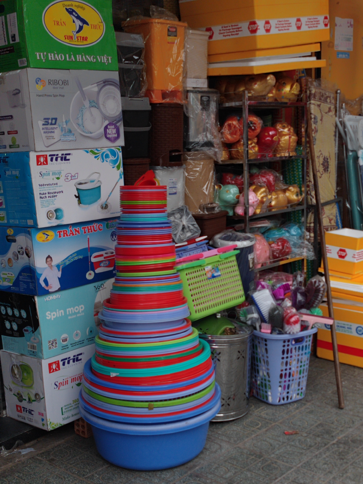 This image captures a densely packed outdoor or semi-outdoor retail space, likely a market stall or storefront in Da Lat, Vietnam, specializing in household goods. The scene is dominated by a tall, tapering stack of vibrant, multi-colored plastic bowls, basins, and plates in the foreground, featuring shades of blue, red, green, and teal.
To the left, a vertical stack of cardboard boxes advertises various "Spin Mop" products. The top green box prominently displays "Doanh nghiệp TRẦN THỨC SUNSTAR" and "TỰ HÀO LÀ HÀNG VIỆT" (Proud to be Vietnamese Goods), accompanied by a shark logo. Below it, a white box for "RIBOBI Hand Press Spin Mop" highlights "3D INTELLIGENT WASHING" and "Brand NEW Upgraded." Other boxes from brands like "THC CLEANING" and "HOMMY" also promote "Spin mop" systems, often mentioning "360 rotated" features and showing images of mop buckets and even a woman demonstrating usage.
On the right side of the image, metal shelving units are filled with a variety of merchandise. Notable items include several golden pig figurines, red decorative pieces, and numerous plastic-wrapped goods. Below the shelves, a blue plastic basket is overflowing with cleaning brushes and other small household items, next to a silver metal bin with patterned cut-outs. Above the shelves, yellow boxes are stacked, bearing "STOP" labels and text indicating "If Seal is Broken Or Package Damaged Check Contents Before Signing Receipt." A long wooden stick leans against the shelving unit on the far right. The ground is paved with patterned grey tiles. No people are visible, and no specific activity is taking place, presenting a static yet vivid display of goods.