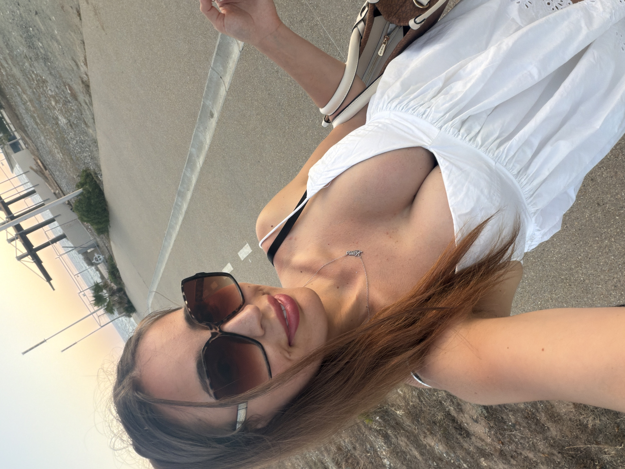 A female subject takes a selfie outdoors in Pervolia, Cyprus, during what appears to be sunset or sunrise, evidenced by the warm-toned sky. She has long, straight brown hair, wears large, square, tortoiseshell-frame sunglasses, and pink lipstick. She is smiling faintly. Her attire consists of a white dress with a deep V-neckline, revealing cleavage, and a visible black bra strap on her left shoulder. A silver necklace spelling "Mama" hangs around her neck. Her right arm is bent, holding a light-colored handbag with brown accents. She stands on a light-colored paved pathway, which features a concrete drainage channel along its left edge. To the left of the path, dry, sparse vegetation is visible. In the background, a body of water (the sea) is visible under a gradient sky of orange, pink, and yellow hues. Structures resembling fencing, beams, and a building are present near the coastline, along with a small palm-like bush.
