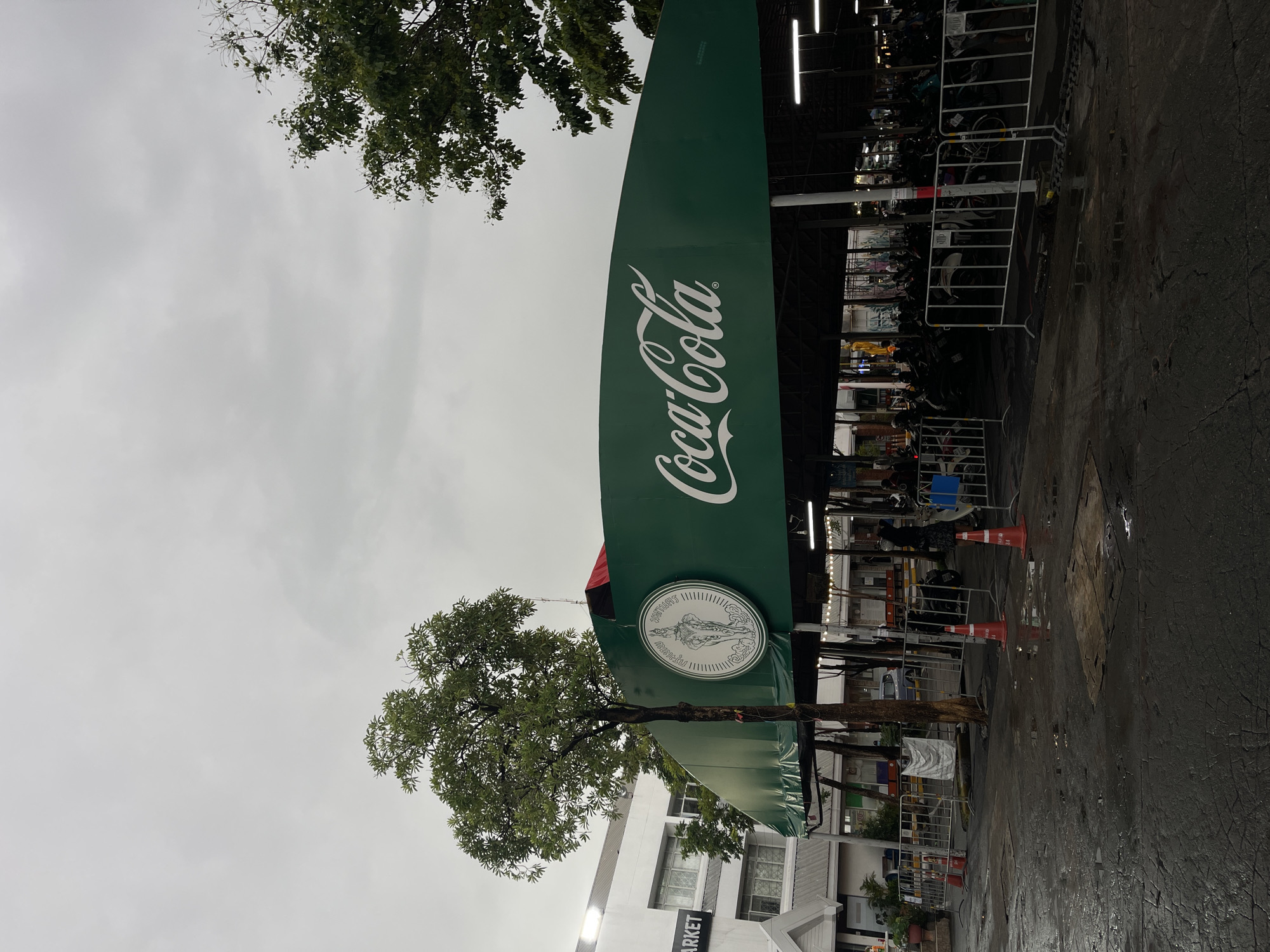 The image shows an outdoor parking area under a large green awning with the Coca-Cola logo prominently displayed on the awning. The awning covers a covered area with many motorcycles parked inside. The ground is wet and reflective, suggesting it has recently rained or is raining. There are orange traffic cones and metal barriers in the foreground, demarcating the parking area. A building with the word "MARKET" on it is visible to the left of the awning. The sky is overcast and grey. The scene appears to be a public parking facility in Bangkok, Thailand.
