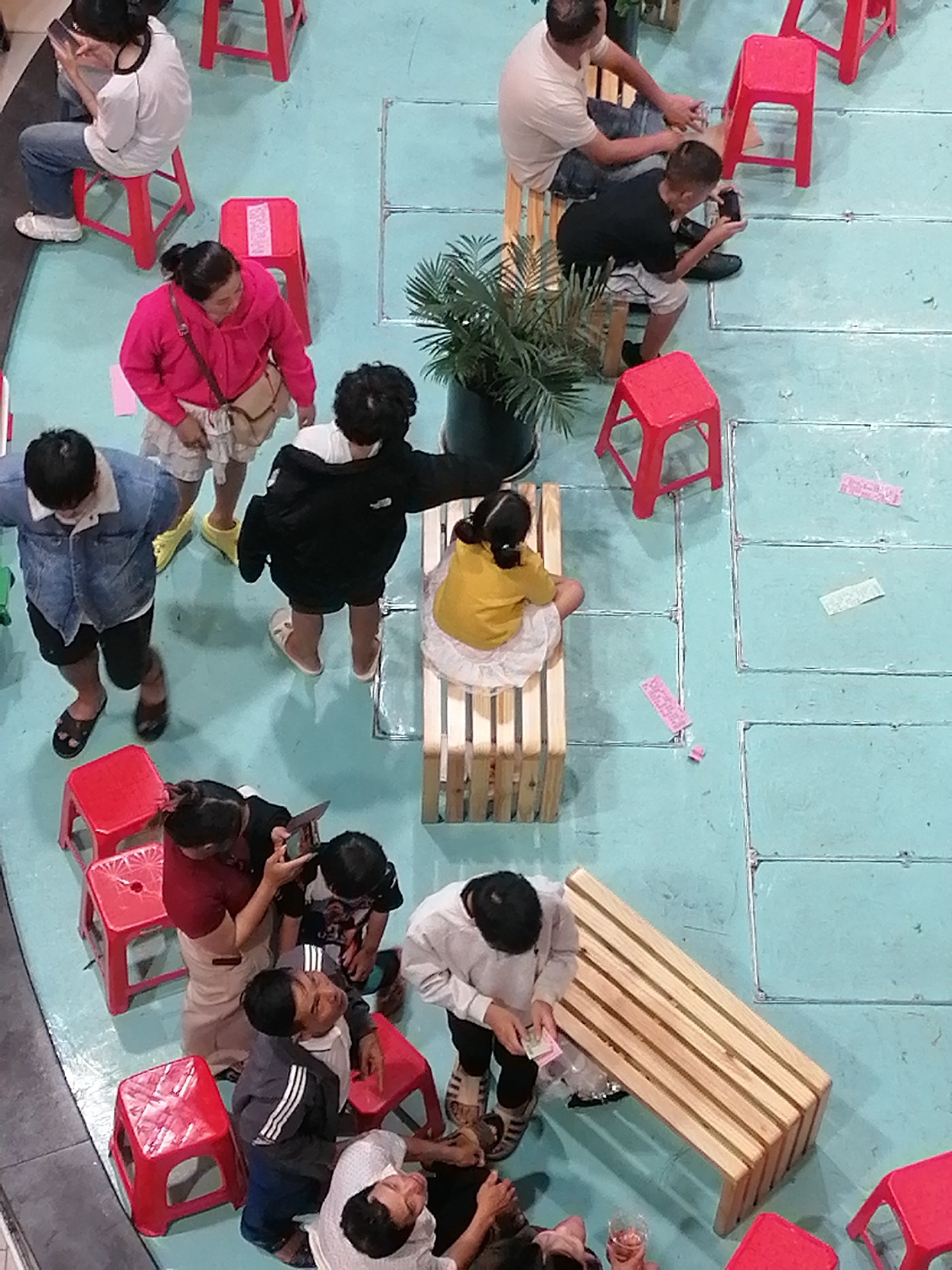 An overhead, eye-level, wide shot captures a group of people gathered in a public space with a light teal floor. In the upper left corner, a woman in a white shirt and jeans is seated on a red stool, engrossed in her phone. Next to her, a woman in a bright pink hoodie and a skirt stands, also looking down. In the upper center, a man in a light-colored t-shirt sits on a wooden bench, while a boy in a black t-shirt and shorts is seated on a red stool, looking at his phone. To the right, a girl in a yellow shirt and white skirt is seated on a wooden bench, facing away from the camera. In the middle left, a person in a denim jacket and shorts stands, looking down. Beside them, a person in a black jacket and shorts is standing, facing right. In the lower left, a child in a red top is seated on a red stool, holding a phone. To the right of the child, two adults are interacting with them. Further to the right, a group of adults are gathered around a wooden bench, with one person in a grey jacket and another in a white shirt seated on the floor. The environment appears to be an indoor public area, possibly a recreational space or waiting area, with various red stools and wooden benches interspersed on the teal floor. The scene is devoid of any specific landmarks or architectural elements that would definitively place it in Da Lat, Vietnam, or any other specific city.