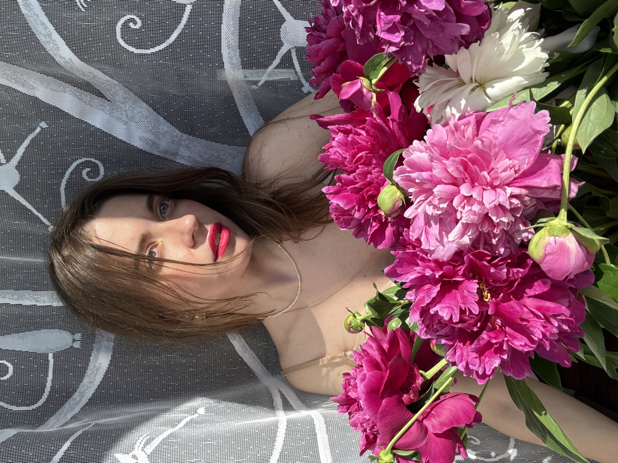 A young woman with brown hair and blue eyes, wearing bright red lipstick, is partially obscured by a bouquet of vibrant pink and white peonies. She is positioned in front of a white lace curtain, through which sunlight is casting shadows. The woman is wearing a beige bra and a delicate pearl necklace. The overall scene appears to be a portrait shot, possibly taken indoors with natural light.