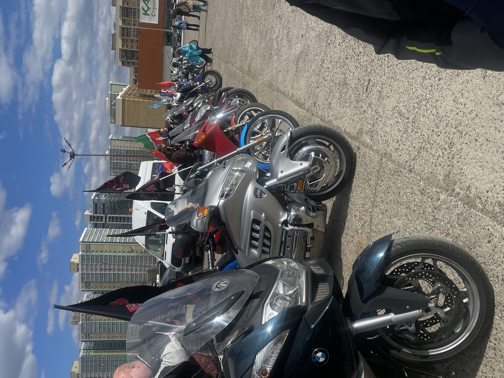 The image shows a row of parked motorcycles, with high-rise apartment buildings visible in the background, which indicates an urban setting. The motorcycles are of different models, colors, and styles, and some have flags attached. A white van is parked among the motorcycles. There are some people in the background. The setting appears to be an open, paved area. The presence of apartment buildings in the background and the motorcycles with flags suggests a gathering or rally. Based on the buildings and general environment, the location is Naberezhnye Chelny, Russia.