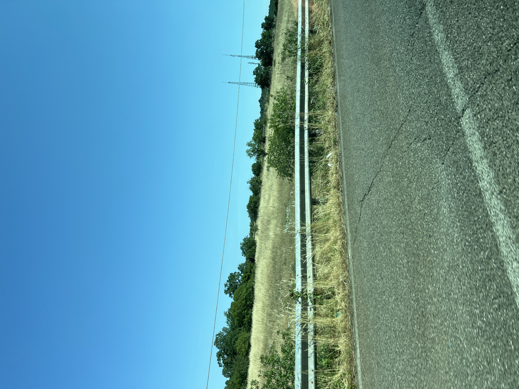 The image displays a roadside scene in Cassano delle Murge, Italy, under a clear, bright blue sky. The upper third of the frame is dominated by the unobstructed blue sky. Below the horizon, a distant line of dark green trees is visible, with a gentle slope of dry, golden-brown grass and scattered green shrubs extending into the midground. Two tall, lattice-structured communication or utility towers stand on the right side of the frame, rising above the tree line. A thin, dark utility wire traverses horizontally across the upper midground.
In the lower midground, a two-tiered silver metal guardrail extends diagonally from left-center to right-center, separating the grassy area from the paved road. The ground immediately adjacent to and behind the guardrail is overgrown with dry grass and weeds, and a piece of white debris is visible near the guardrail on the right side. The lower section of the image reveals a dark asphalt road surface, exhibiting visible cracks and featuring a faded white solid line marking its right edge. The road and guardrail show slight motion blur, consistent with an image captured from a moving vehicle. The scene is devoid of any individuals, vehicles, or animals.