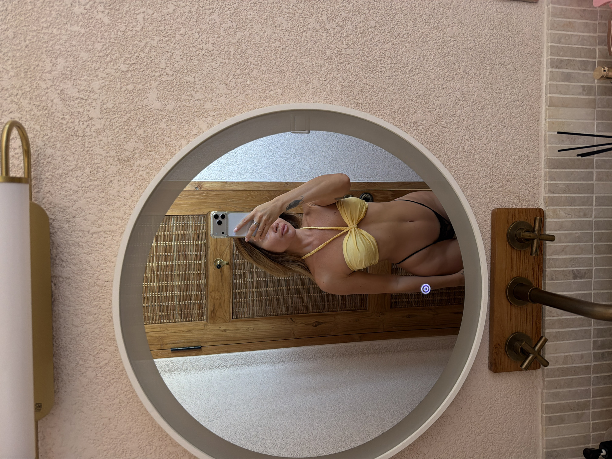 The image shows a woman taking a mirror selfie in a bathroom. She is wearing a yellow bikini top and a black thong. The bathroom has a textured pink wall, a round mirror with a light, and a wooden cabinet behind her. There are also some faucets and tiles visible in the foreground. The woman is holding a smartphone to take the picture. The image does not provide any information about the location in Mengwi, Indonesia.