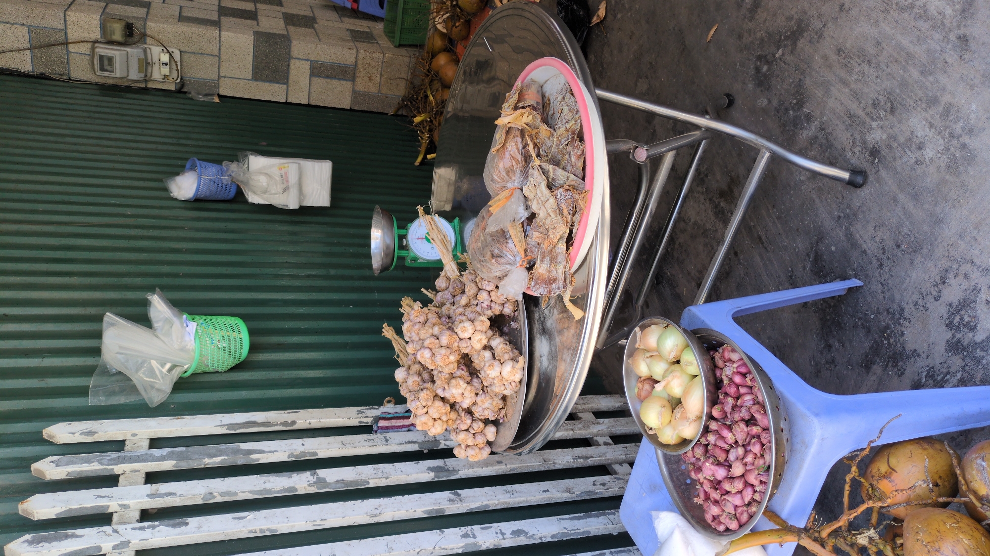 The scene captures an outdoor marketplace in Da Lat, Vietnam, likely a makeshift roadside setup. There are no people visible in the frame.
The primary subjects are food items for sale: large bunches of garlic, bags of dried bamboo shoots, and a bowl containing both white and red onions. These items are displayed on a large metal table. A green and silver scale sits beside the garlic, suggesting that items are weighed for sale.
The immediate environment consists of a corrugated metal wall, a weathered white bench, a blue plastic chair, and the concrete surface. Coconuts are arranged on the ground near the onions. The setting is simple and functional, indicative of a typical street vending arrangement in Vietnam.