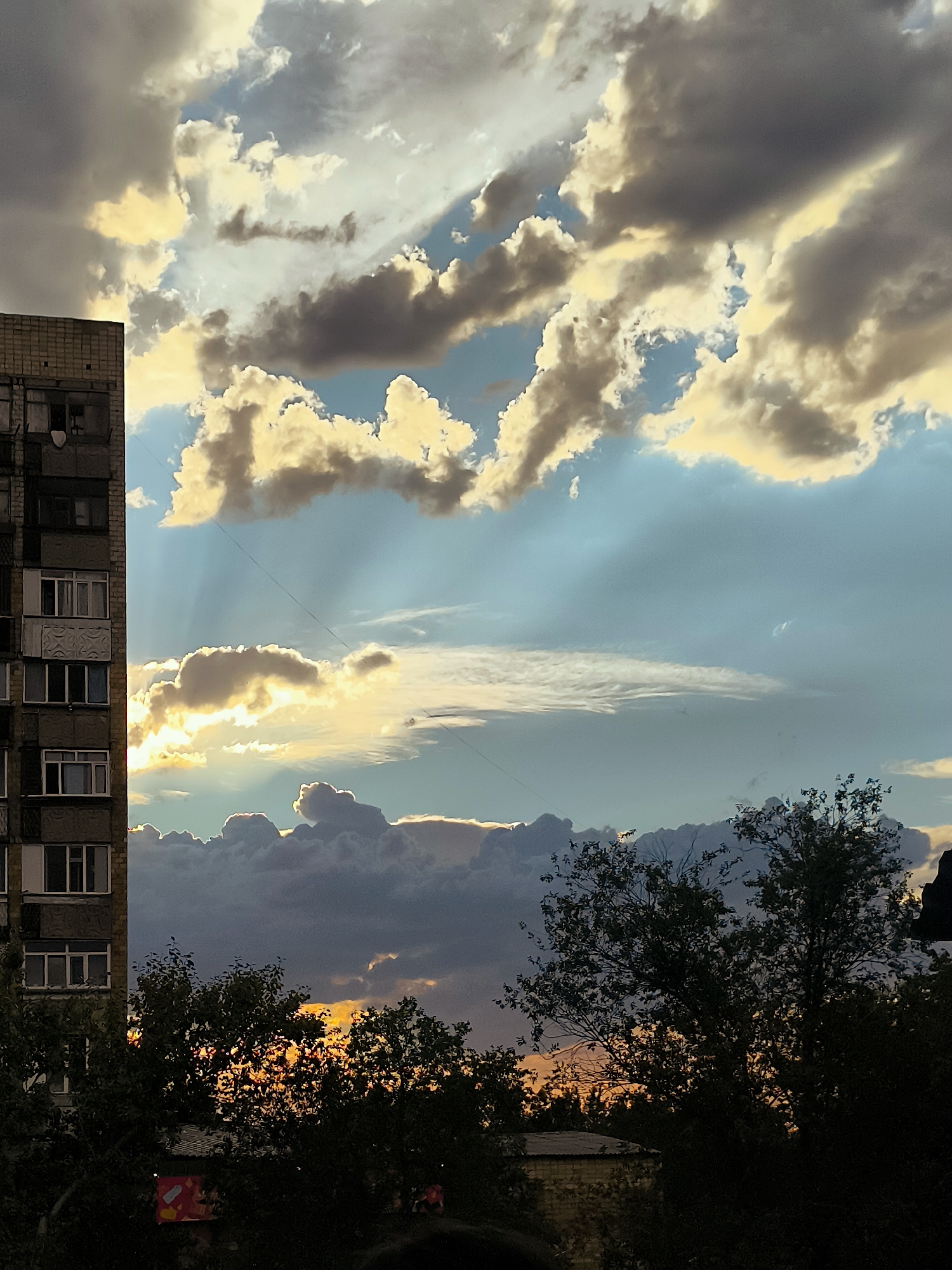 An outdoor, eye-level, vertical shot shows a building and trees against a sky filled with dramatic clouds during sunset. The building, on the left, is a tall, multi-story structure with a brick facade and numerous windows. The sky is a mix of blue, grey, and golden clouds, with shafts of sunlight breaking through. Trees fill the lower portion of the frame, their dark silhouettes contrasting with the bright sky. The overall impression is of a cloudy sunset viewed from an urban environment in Qarağandı city administration, Kazakhstan.
