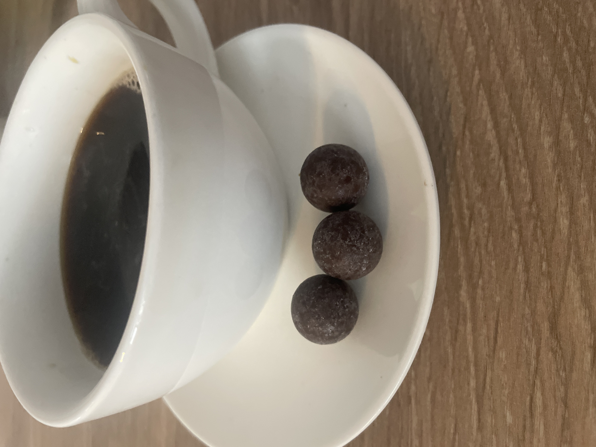A close-up, high-angle shot shows a white coffee cup filled with dark liquid and a white saucer with three dark brown spherical candies on it. The cup and saucer are positioned on a wooden surface. The image appears to have been taken indoors, likely on a table. There are no people visible in the image. The candies are roughly the same size and appear to be made of chocolate. The coffee is dark and appears to have a slight sheen on its surface. The wooden surface has a visible grain pattern. There is no specific information that can place this scene in Brovary, Ukraine.