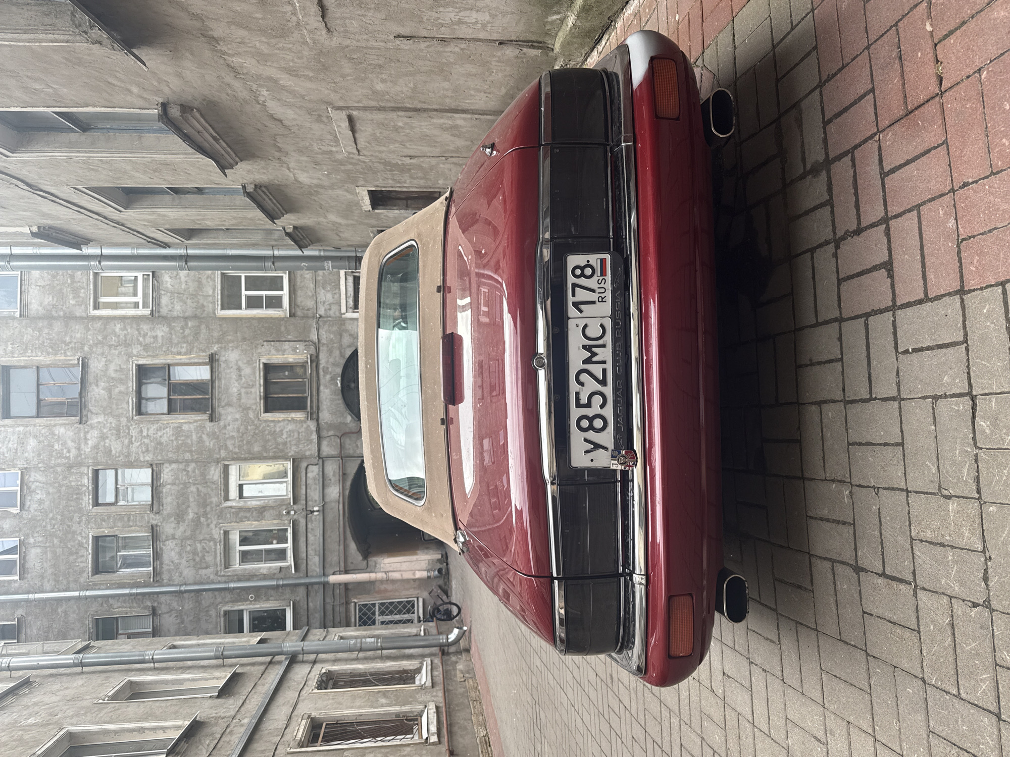 A dark red convertible Jaguar is parked on a brick-paved courtyard in Saint Petersburg, Russia. The car is viewed from the rear, with its trunk and taillights prominently displayed. The license plate reads "y852MC 178 RUS". The courtyard is enclosed by weathered, multi-story buildings with numerous windows and exposed pipes. A bicycle is visible in the background, leaning against a wall. The overall scene suggests an older, historic urban setting.